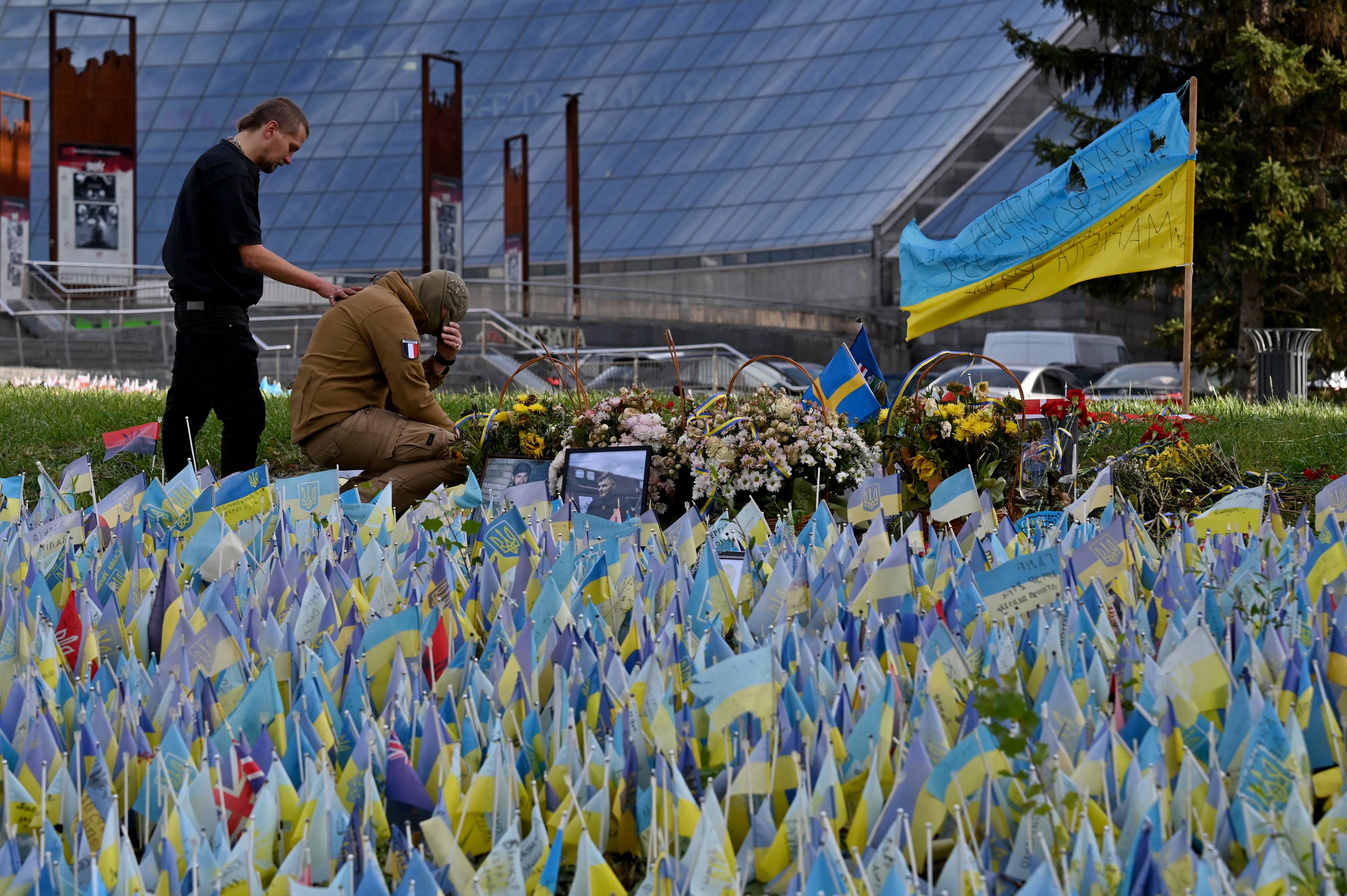 mężczyzna pochyla się nad klęczącym i rozpaczającym kolegą na placu pełnym ukraińskich niebiesko-żółtych chorągiewek