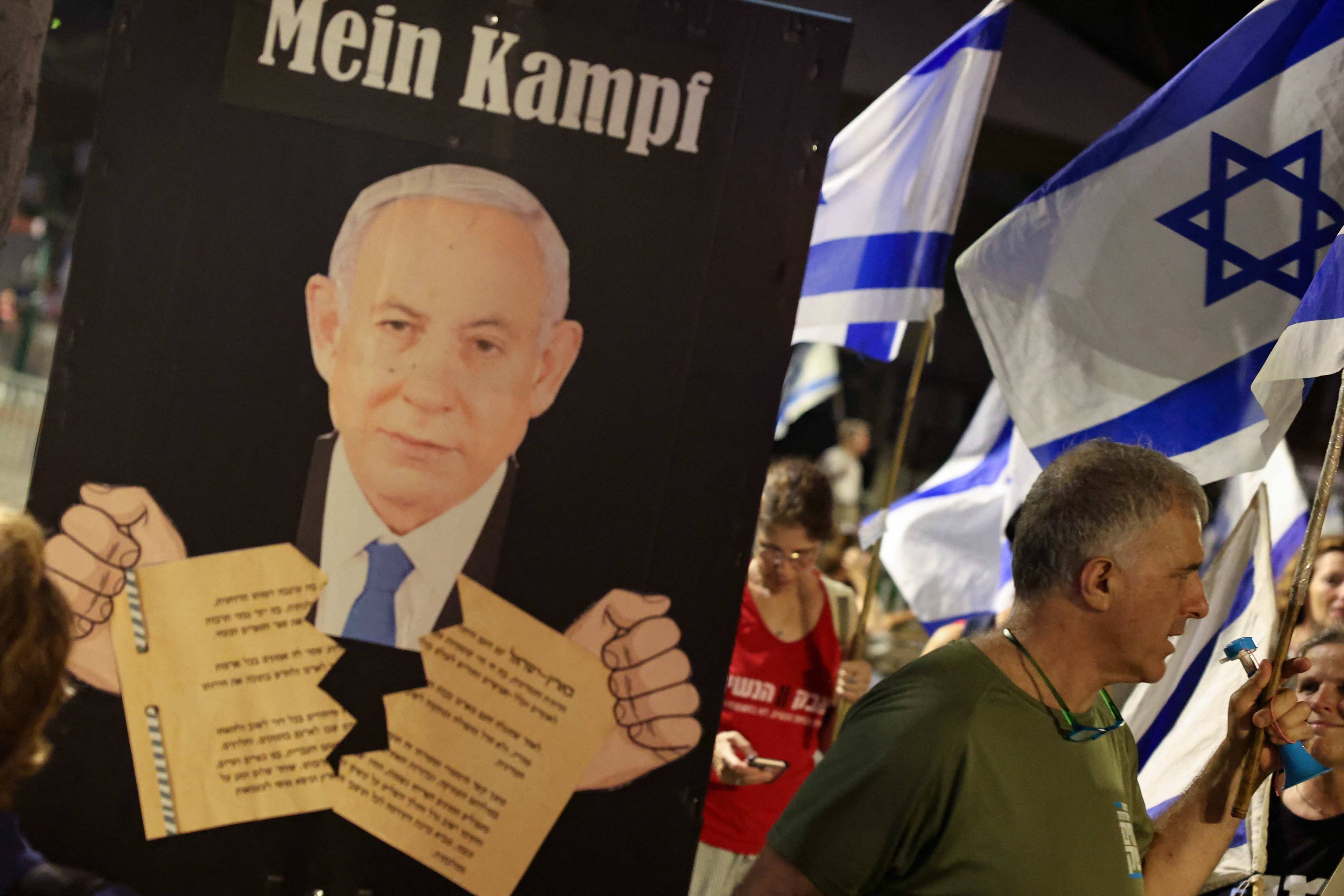 Demonstranci idą ulicą, trzymając w rękach flagi Izraela. Na pierwszym planie plakat z podobizną Benjamina Netanjahu, który rwie w rękach dokument w języku hebrajskim. Nad nim napis Mein Kampf