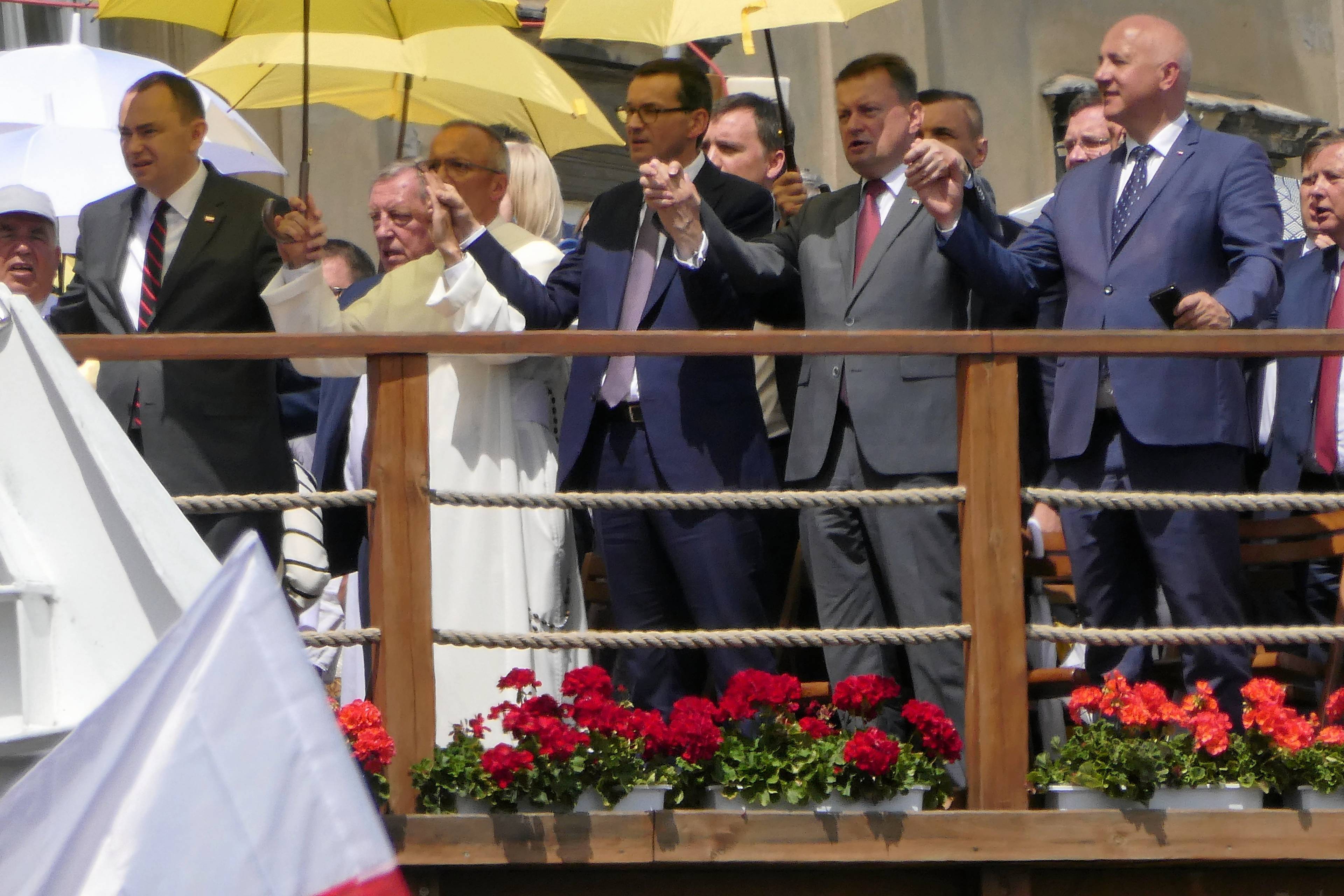 Mateusz Morawiecki, Mariusz Błaszczak i zakonnik w białym habicie trzymają się za ręce, nad nimi żółte parasole