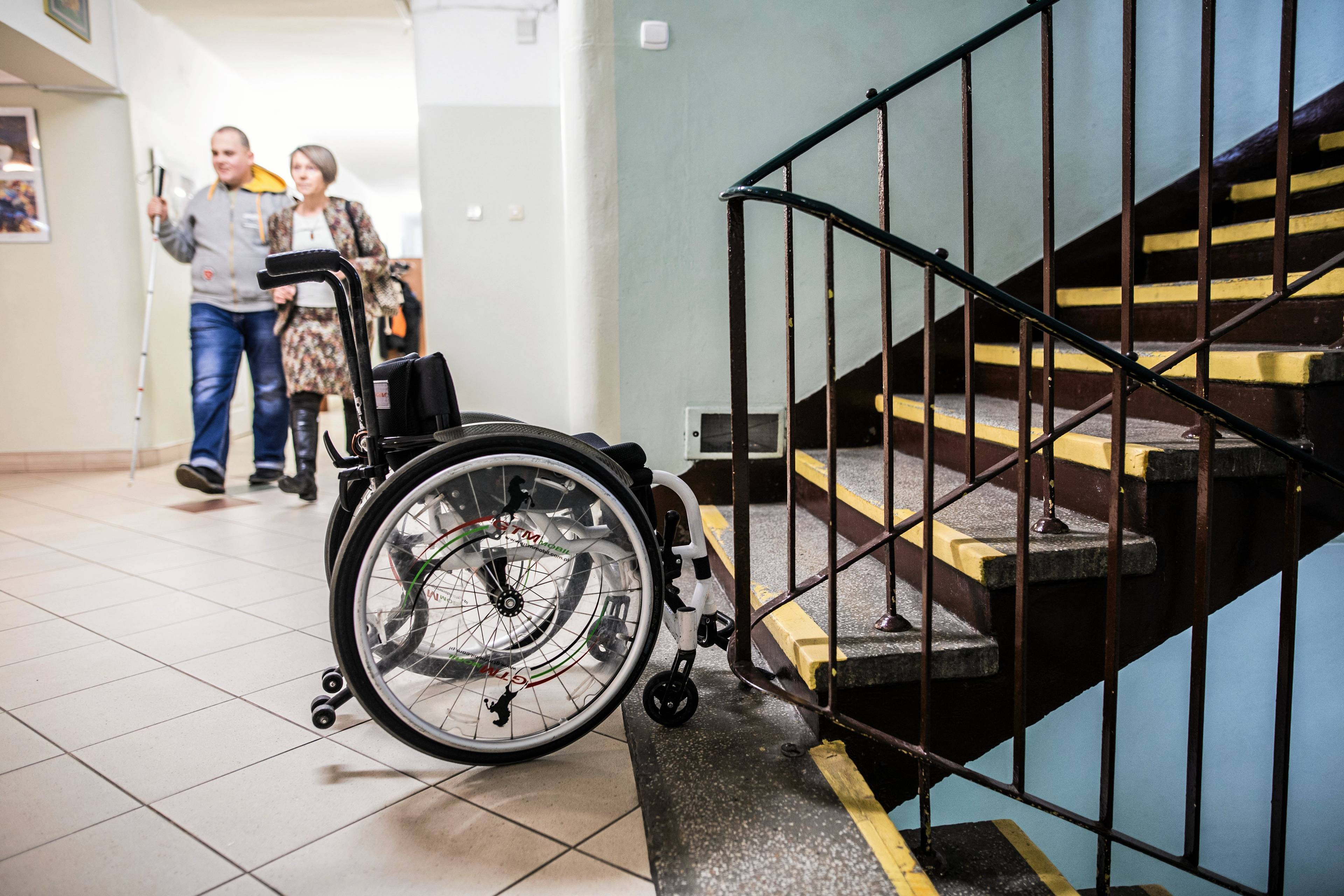 Wózek inwalidzki przed schodami na klatce. W tle - dwoje ludzi idzie korytarzem
