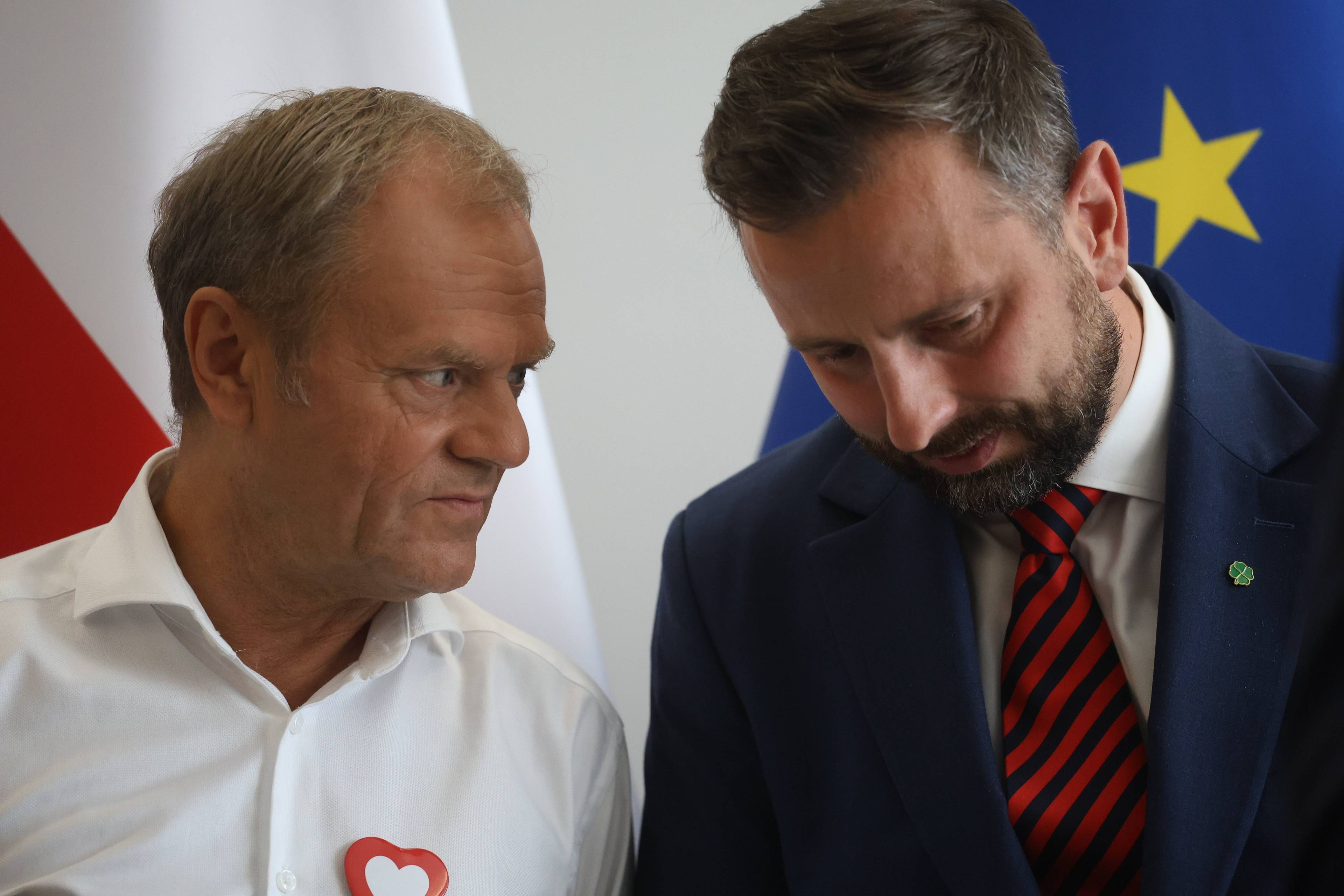 Władysław Kosiniak-Kamysz po lewej z pochyloną głową na tle flagi Unii Europejskiej słucha, co mówi do niego Donald Tusk na tle polskiej flagi