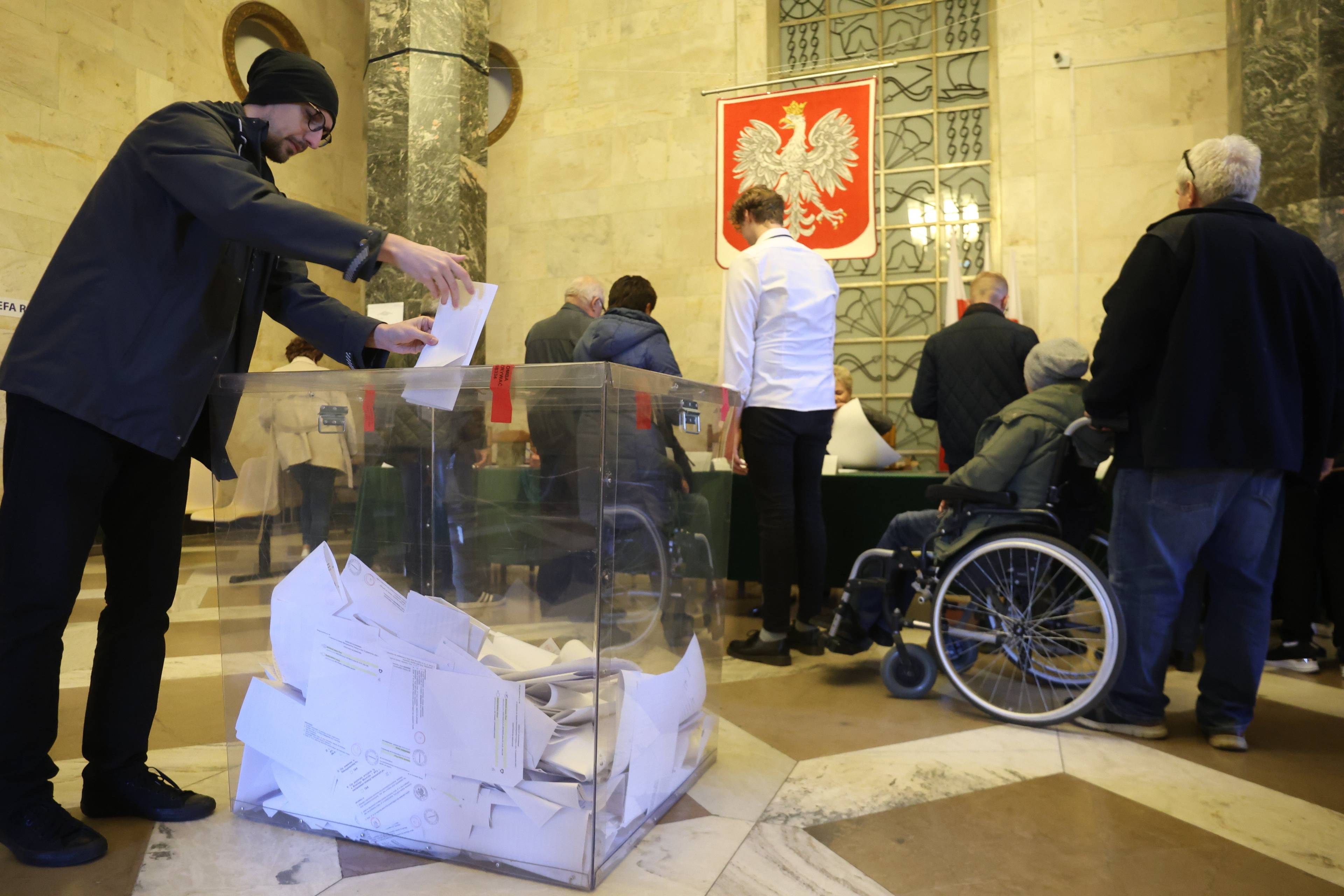 Komisja wyborcza. Mężczyzna wrzucający kartę do urny, w tle kolejka po karty do głosowania, w niej osoba na wózku inwalidzkim