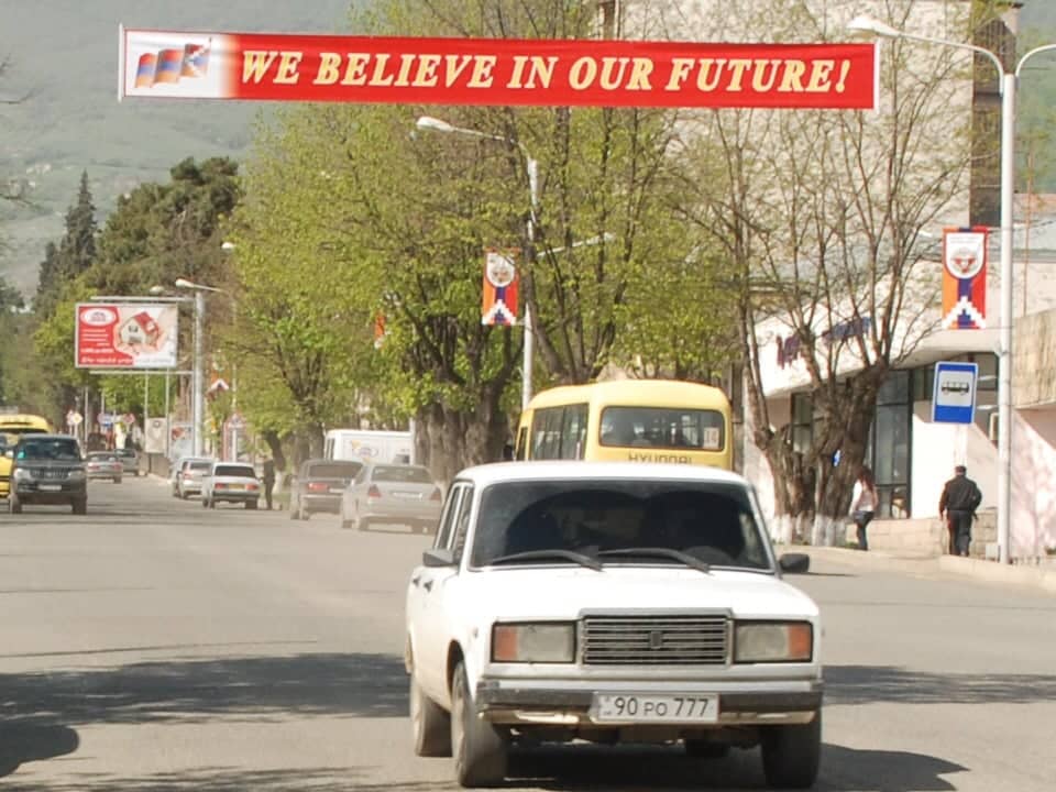 ulica, na pierwszym planie jedzie biała łada, nad ulicą czerwony transparent z napisem po angielsku: wierzymy w naszą przyszłość