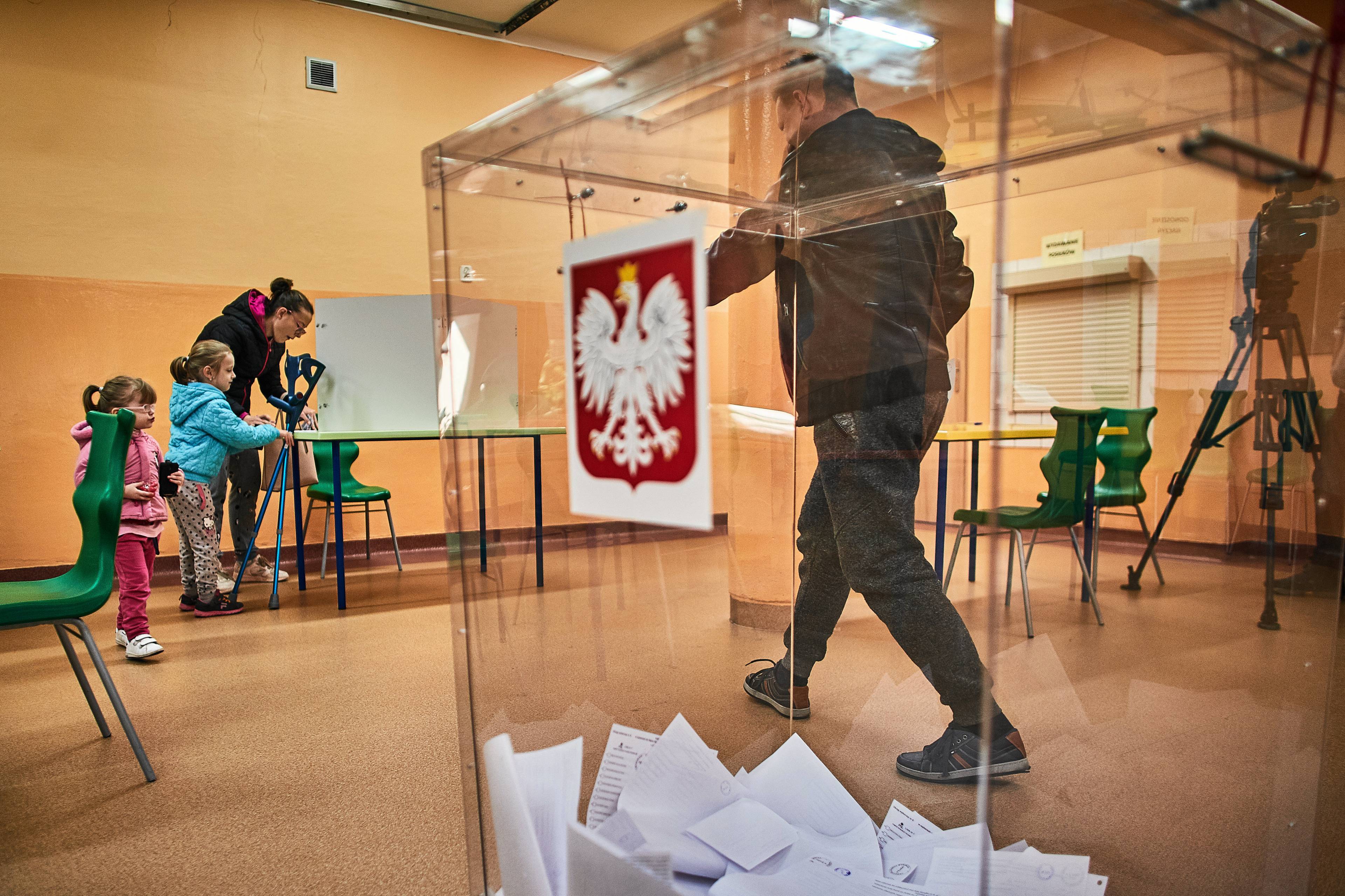 Przezroczysta urna wyborcza z naklejonym polskim godłem, na jej spodzie karty do głosowania, za nią ludzie