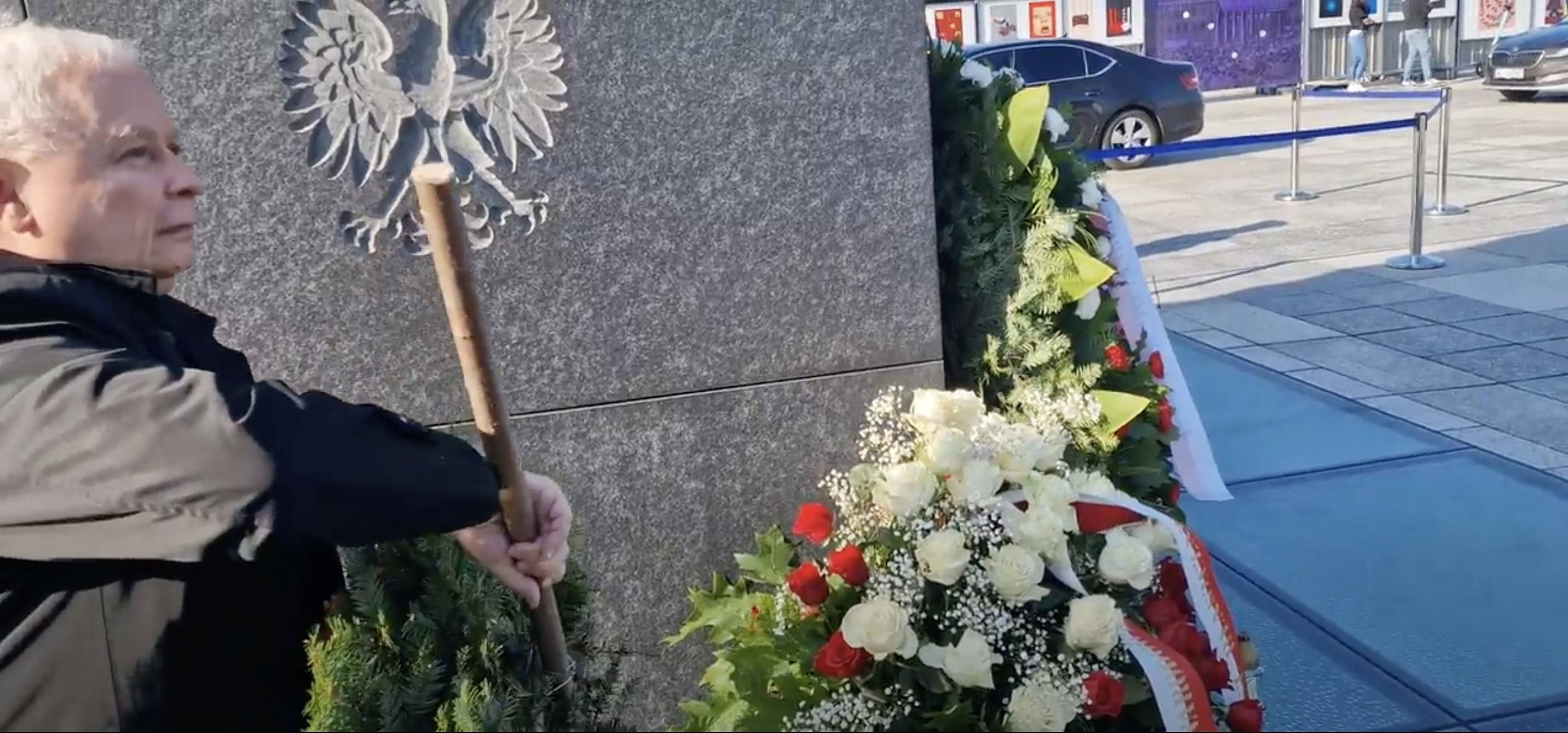 Jarosław Kaczyński trzyma drzewiec wieńca pod pomnikiem ofiar katastrofy smoleńskiej w Warszawie. W tle godło Polski