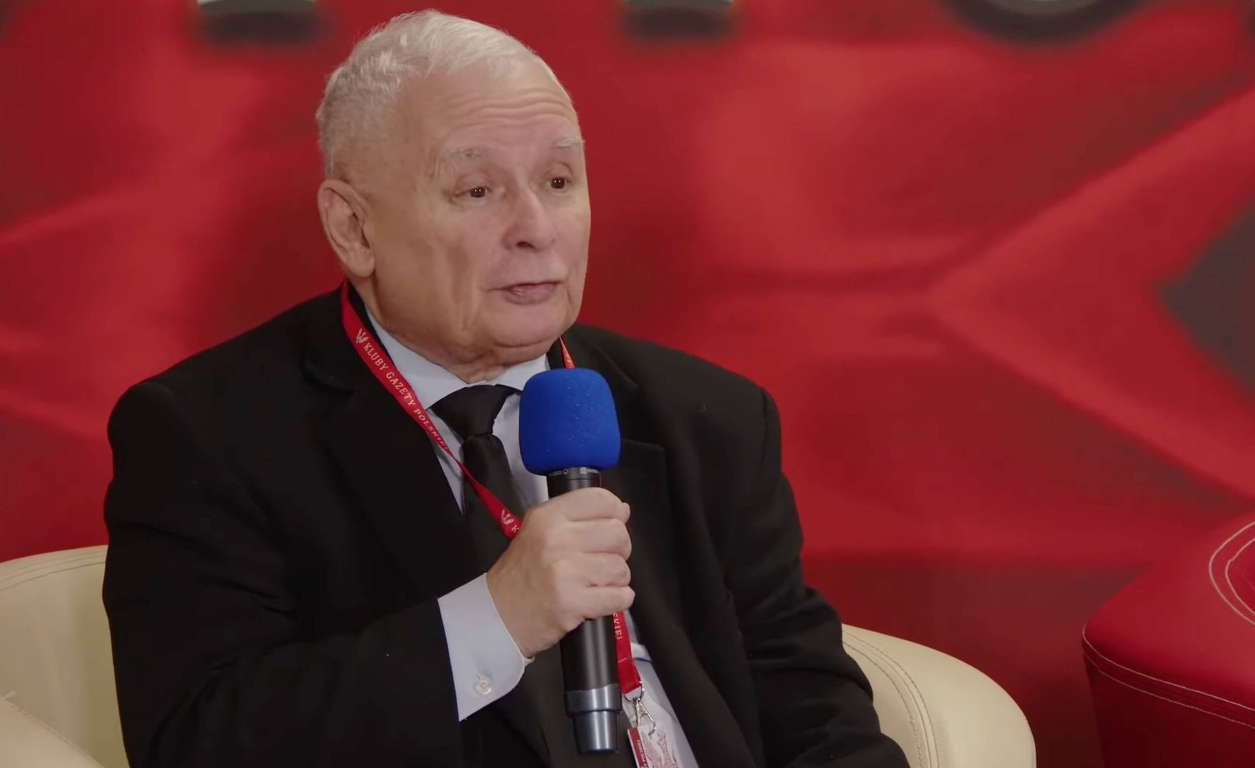 Jarosław Kaczyński mówi do mikrofonu, z tyłu czerwone tło