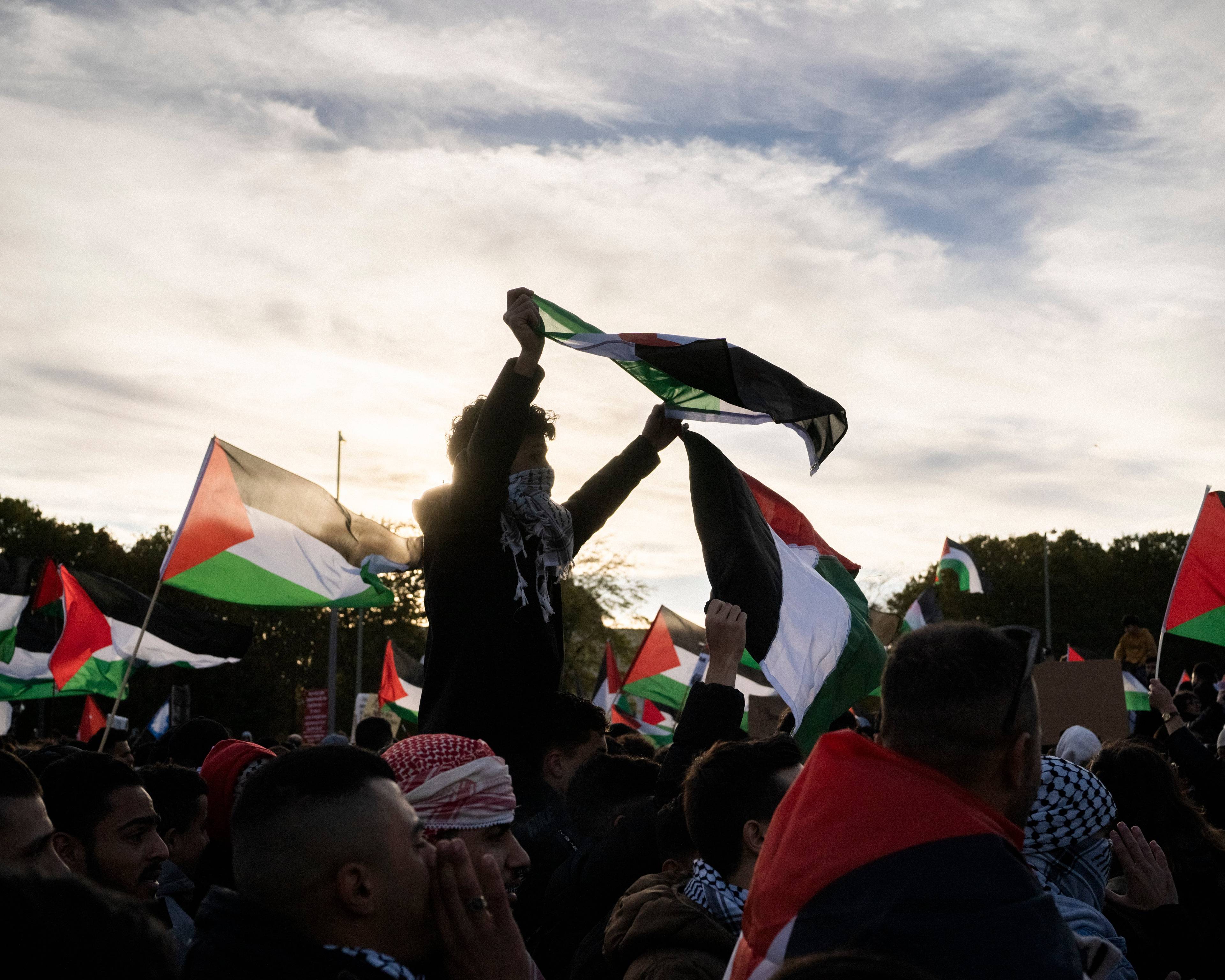chłopak w arafatce na twarzy niesiony na ramionach demonstrantów trzyma flagę czarno-biało-zielono-czerwoną powiewającą na wietrze