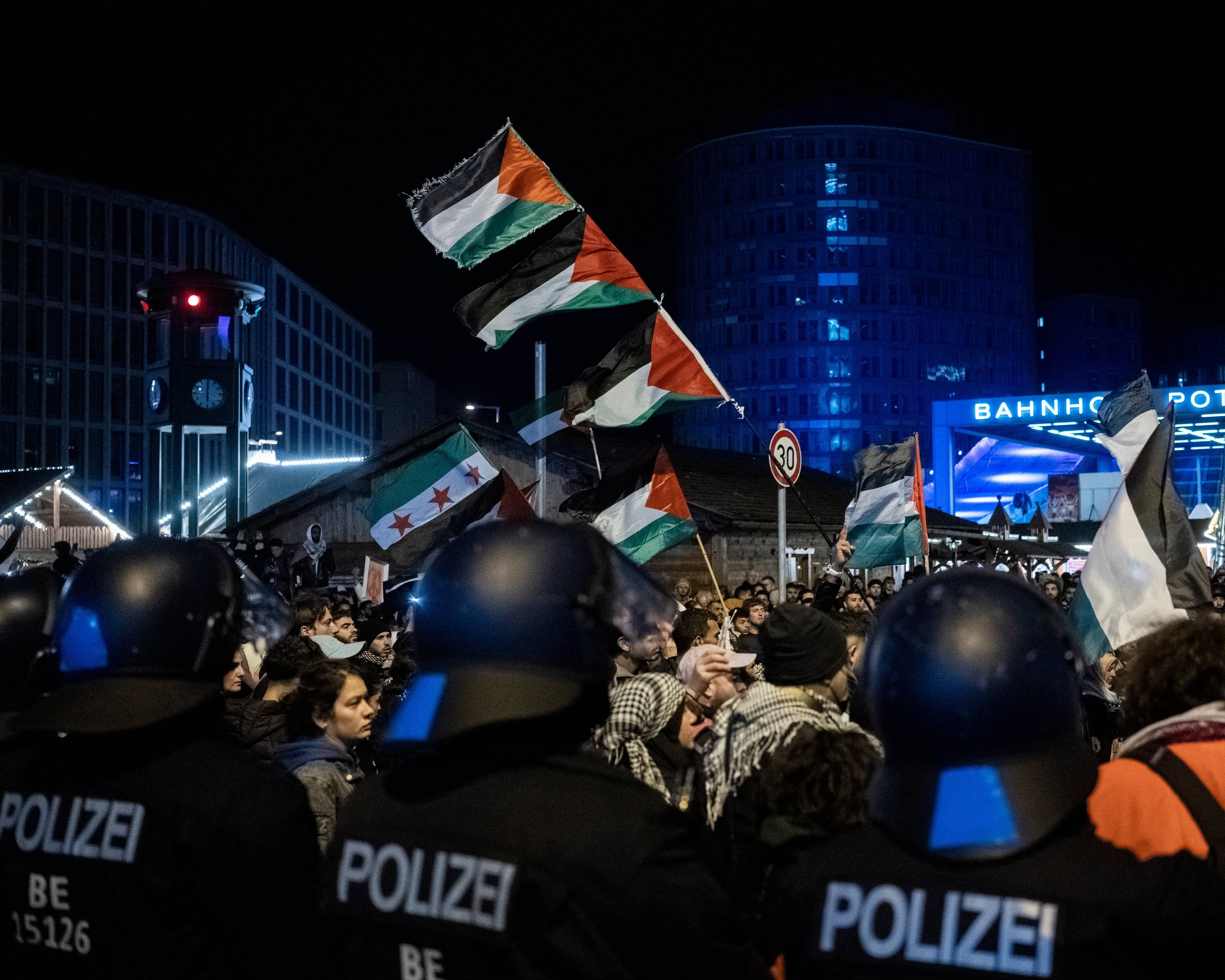 zdjęcie demonstracji wieczorem, na pierwszym planie plecy niemieckich policjantów w kaskach, widać flagi Palestyny i Syrii