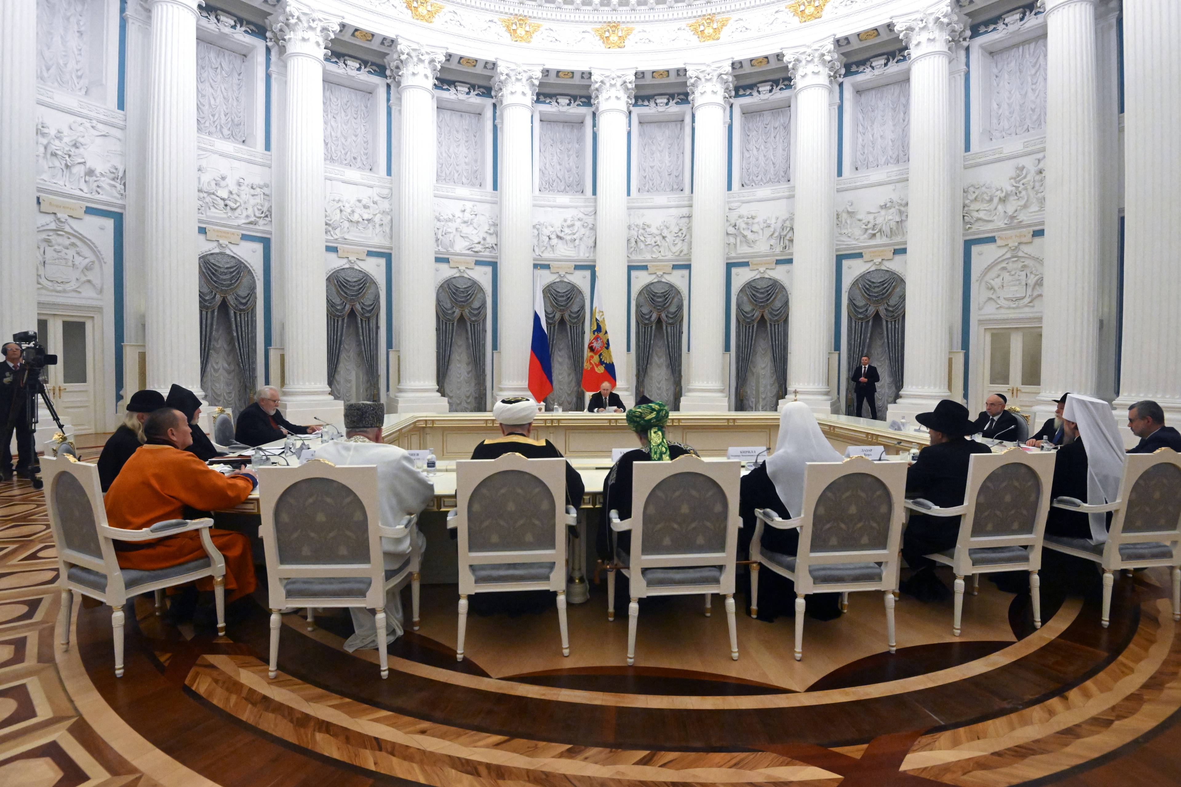 W wielkiej sali z kolumnadą za stołem siedzą dostojnicy religijni a naprzeciwko - Putin
