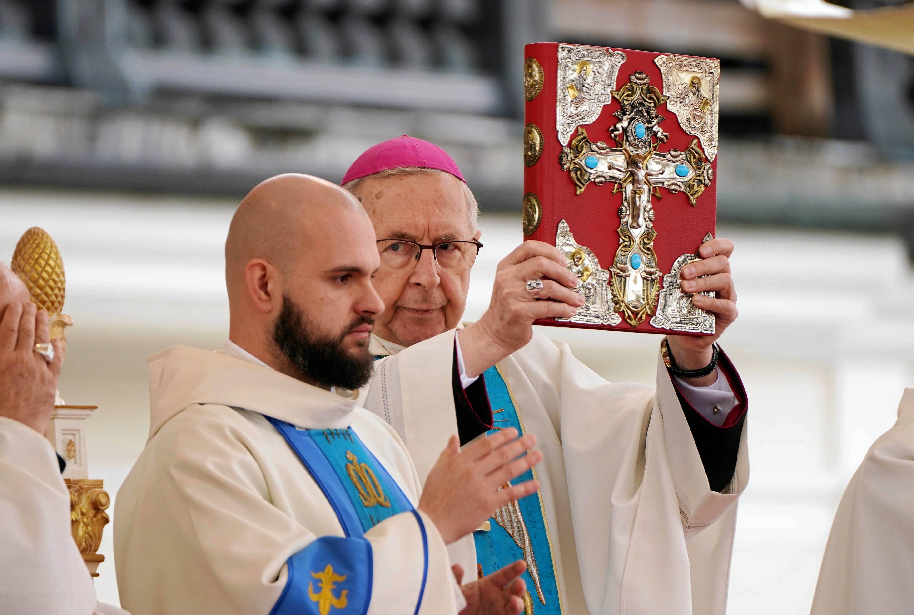 Arcybiskup Stanisław Gądecki trzyma przed sobą bogato zdobioną Biblię ze złotym krzyżem na okładce, obok stoi brodaty zakonnik.