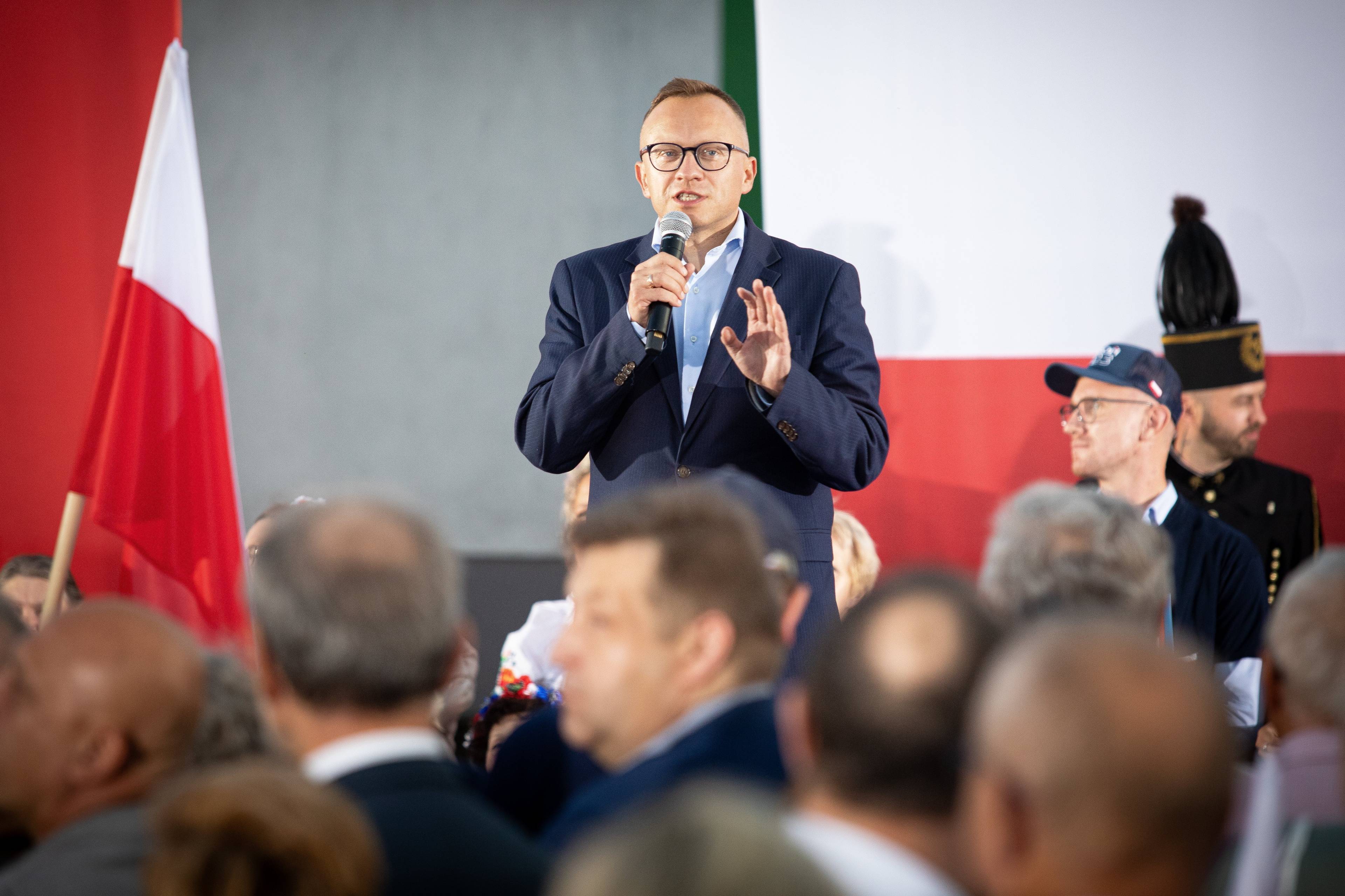 Wiceminister finansów Artur Soboń przemawia do tłumu zgromadzonego w sali na tle białoczerwonej flagi