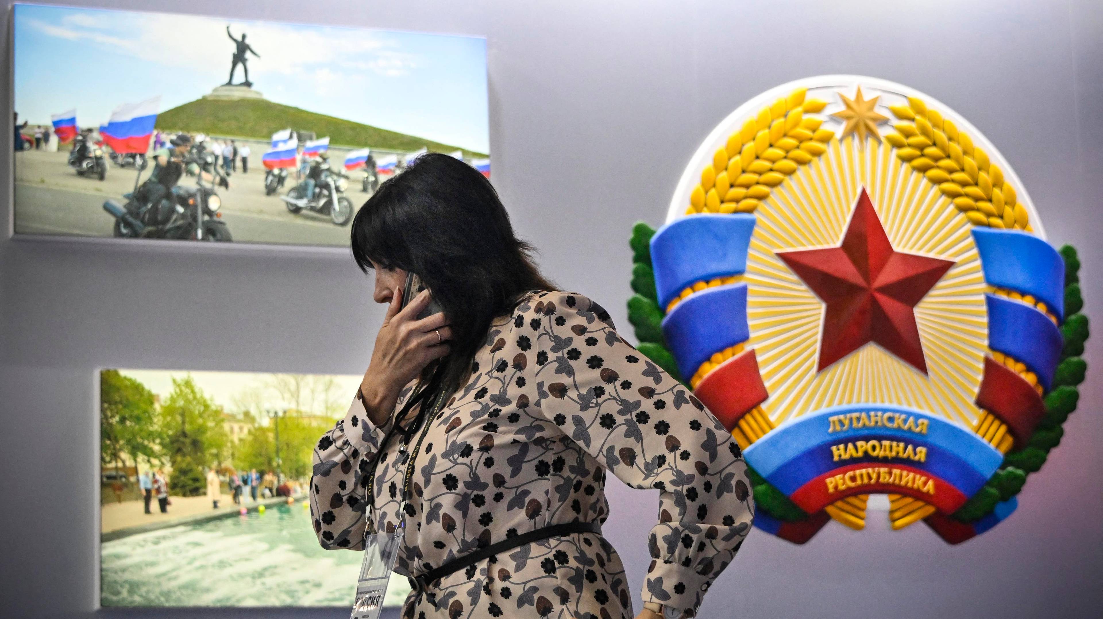 Kobieta rozmawia przez telefon na tle ściany wystawowej, na której wisi godło Ludowej Republiki Ługańskiej z czerwoną gwiazdą oraz zdjecia, przedstawiające rajd motocyklowy i fontannę