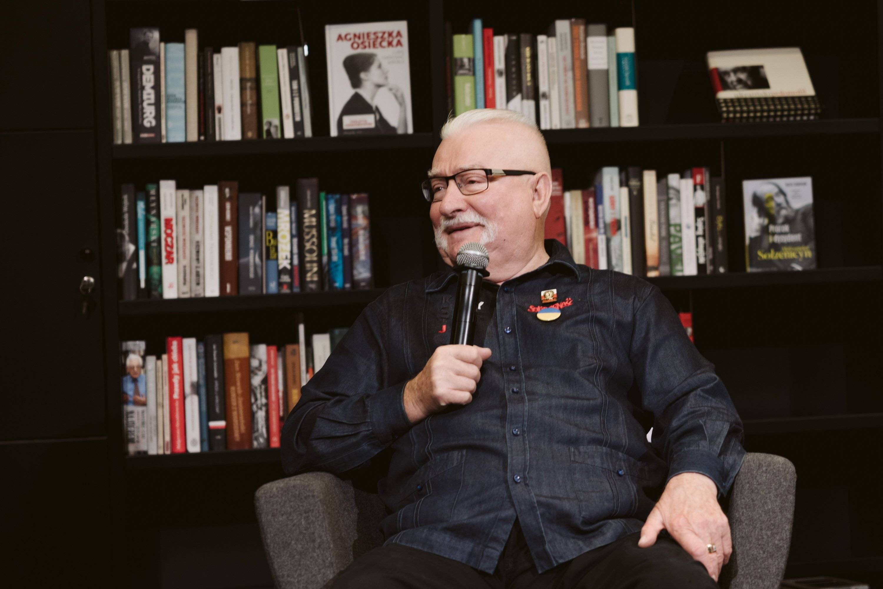 Lech Wałęsa siedzi na tle regału z książkami. Ma na sobie ciemną koszulę, okulary i trzyma mikrofon