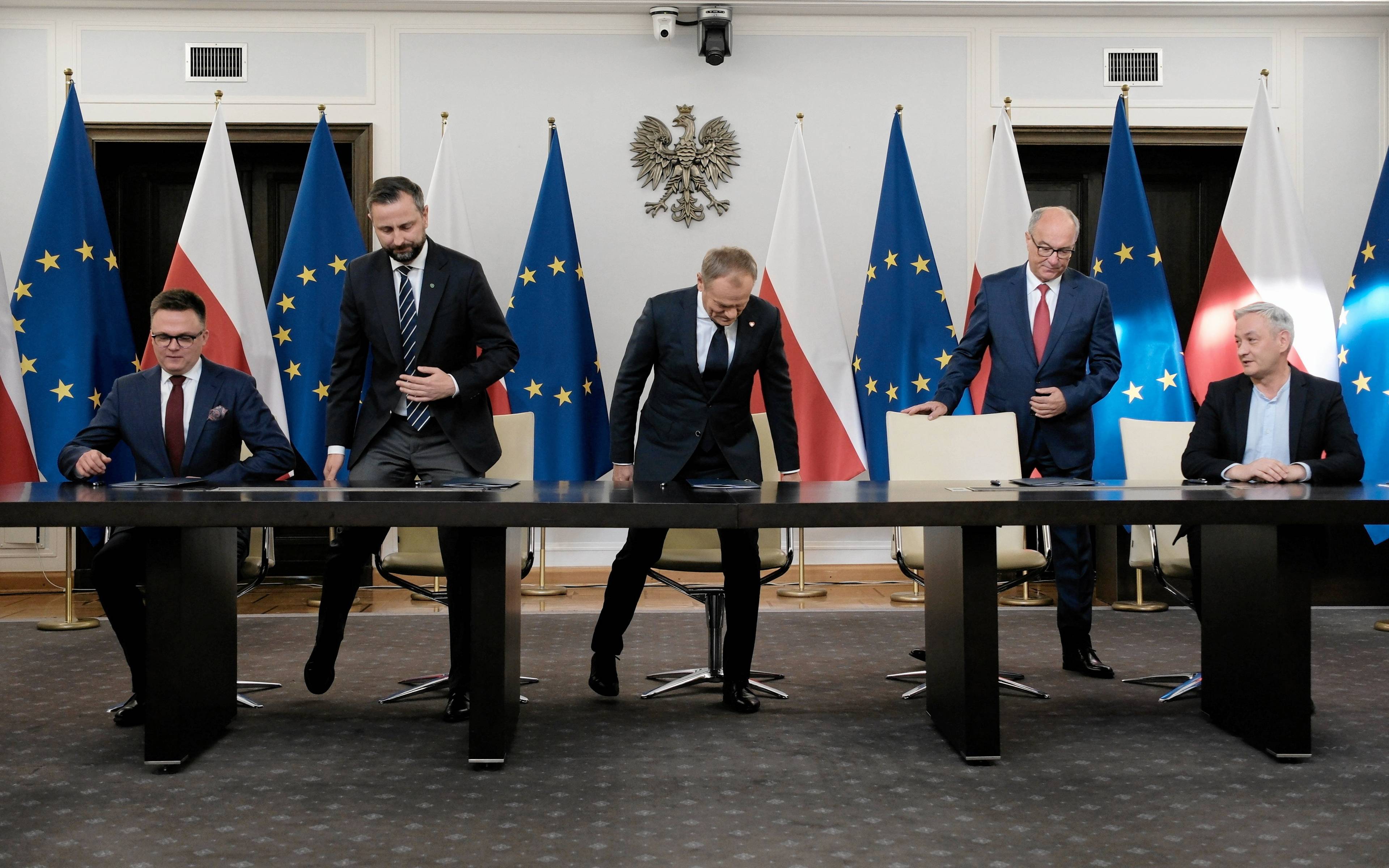 Liderzy nowej większości rządowej wstają od stołu chwilę po podpisaniu umowy koalicyjnej, w tle flagi Polski i UE oraz godło Polski