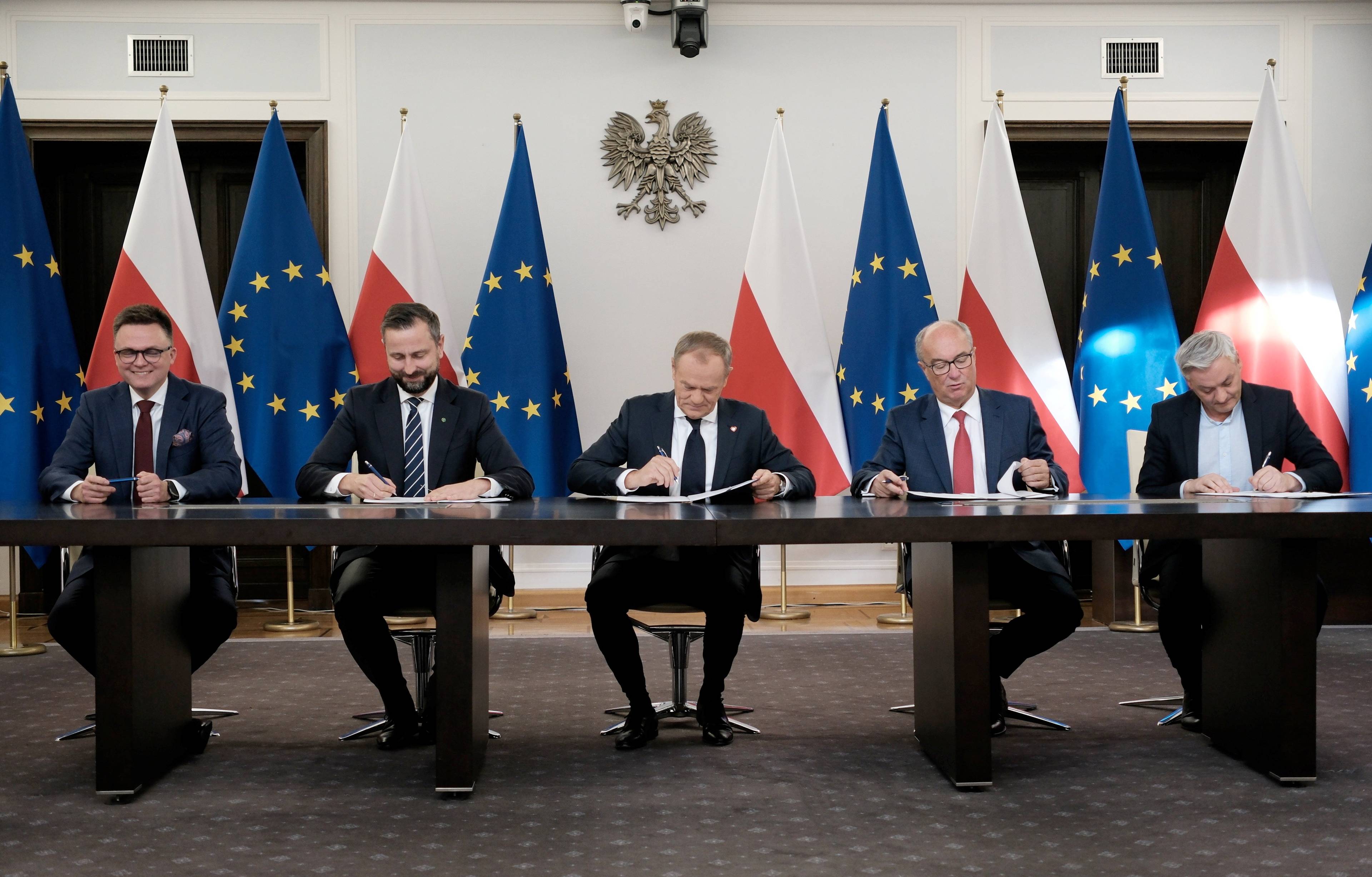 Liderzy KO, Trzeciej Drogi i Nowej Lewicy podpisują przy długim stole umowę koalicyjną, za nimi godło Polski oraz flagi - Polski i Unii Europejskiej