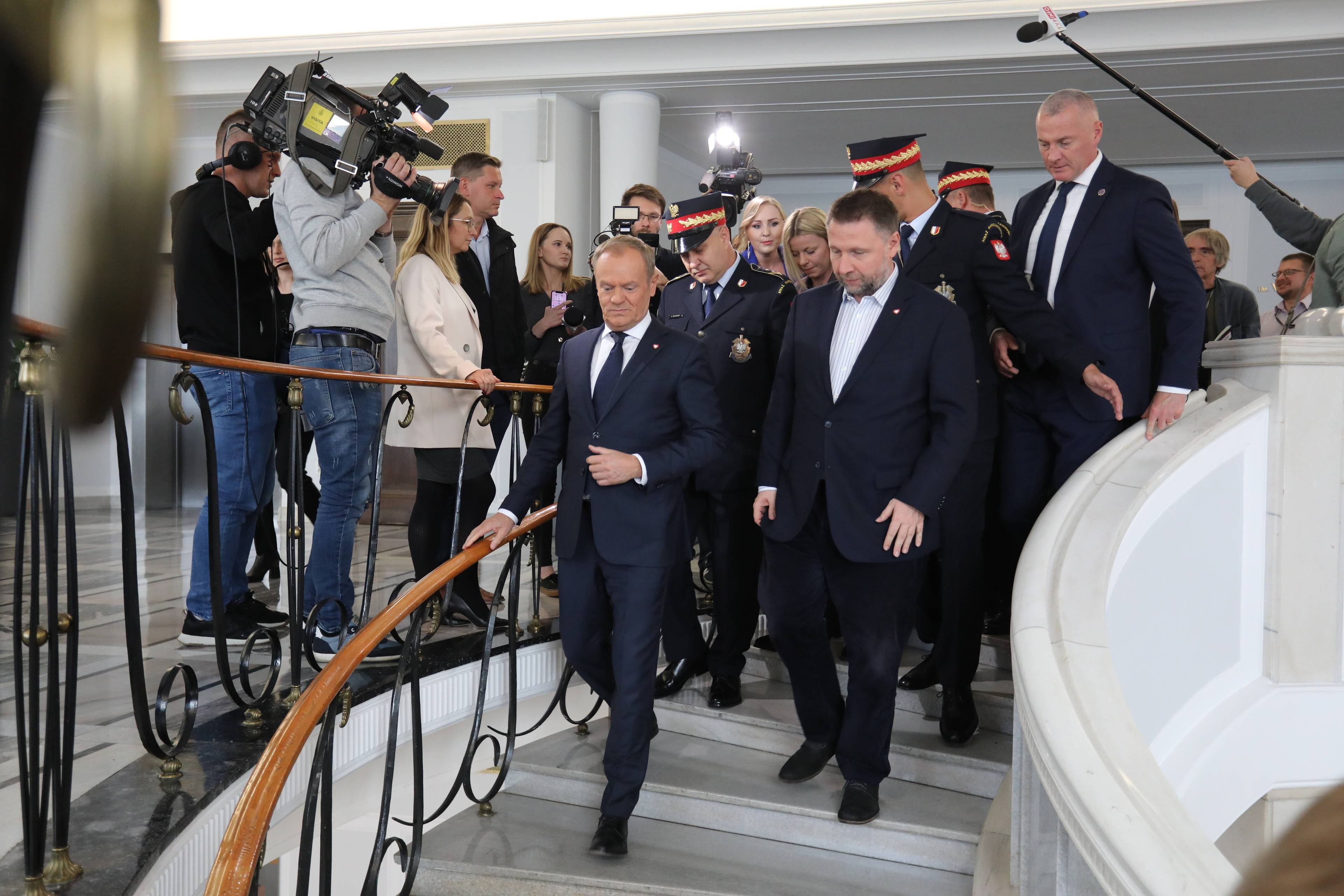 Donald Tusk schodzi po schodach otoczony przez straż marszałkowską. W tle dziennikarze starający się zadać mu pytanie