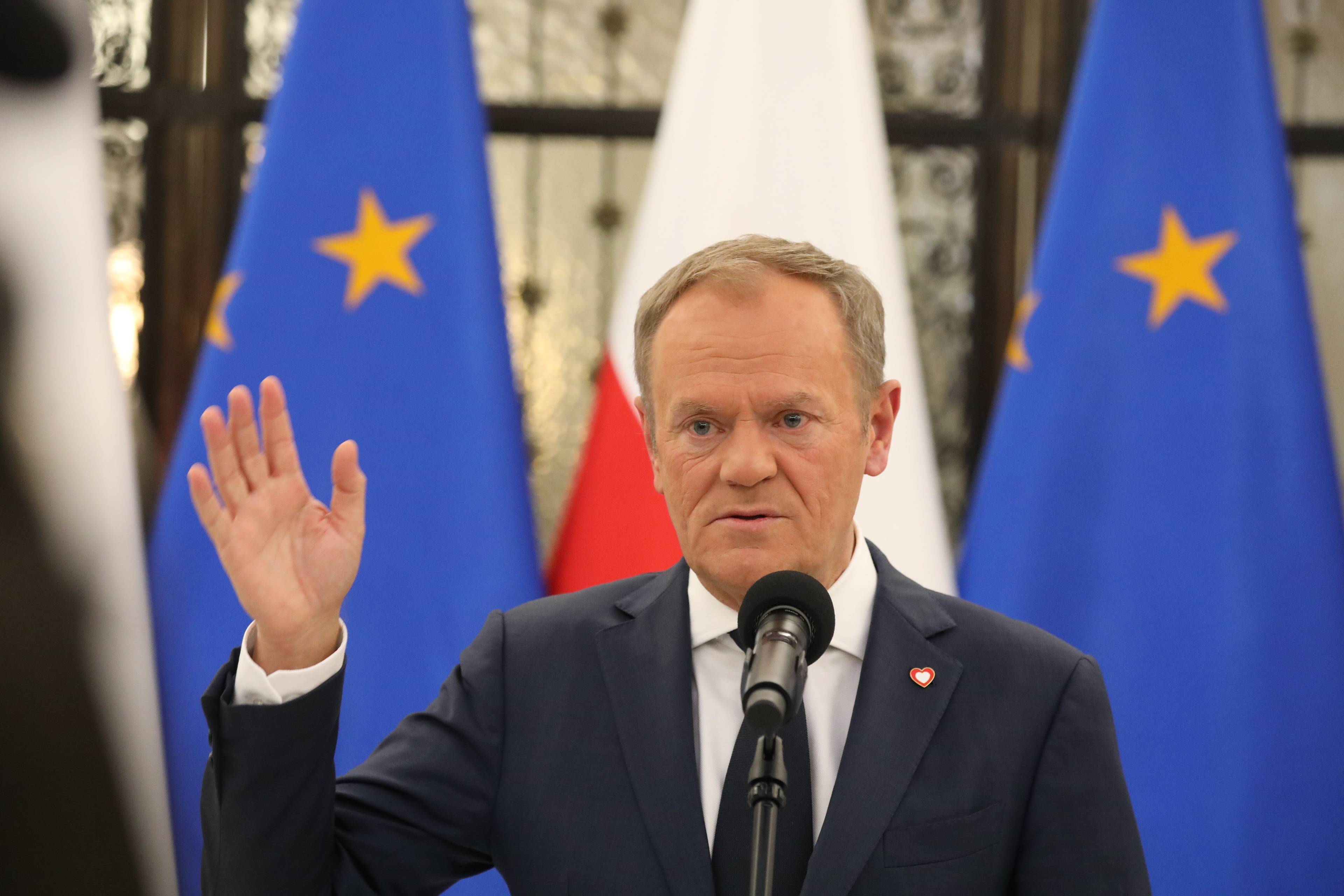 Donald Tusk stoi na tle flagi Polski i Unii Europejskiej, lewą rękę ma wzniesioną do góry