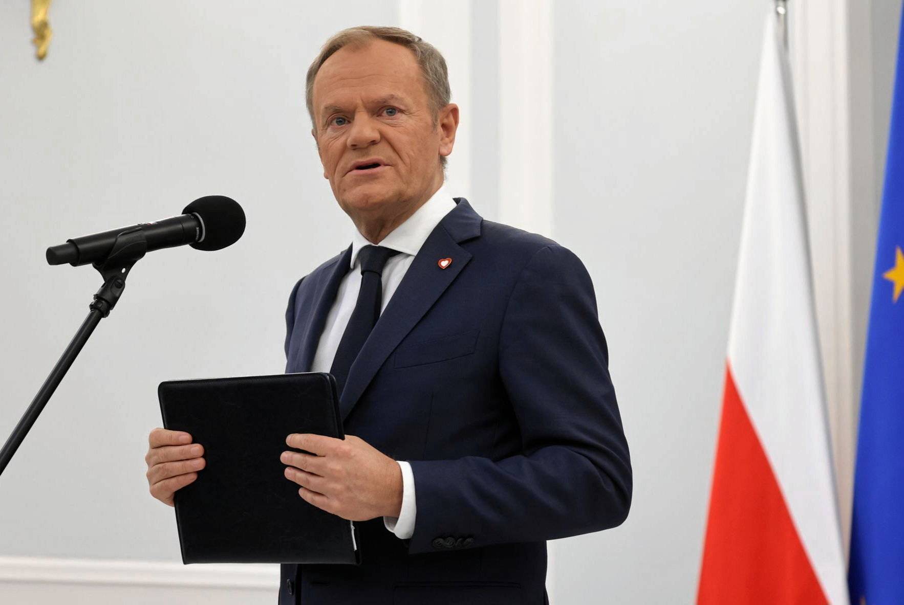 Donald Tusk przemawia przed mikrofonem na tle flag Polski i UE