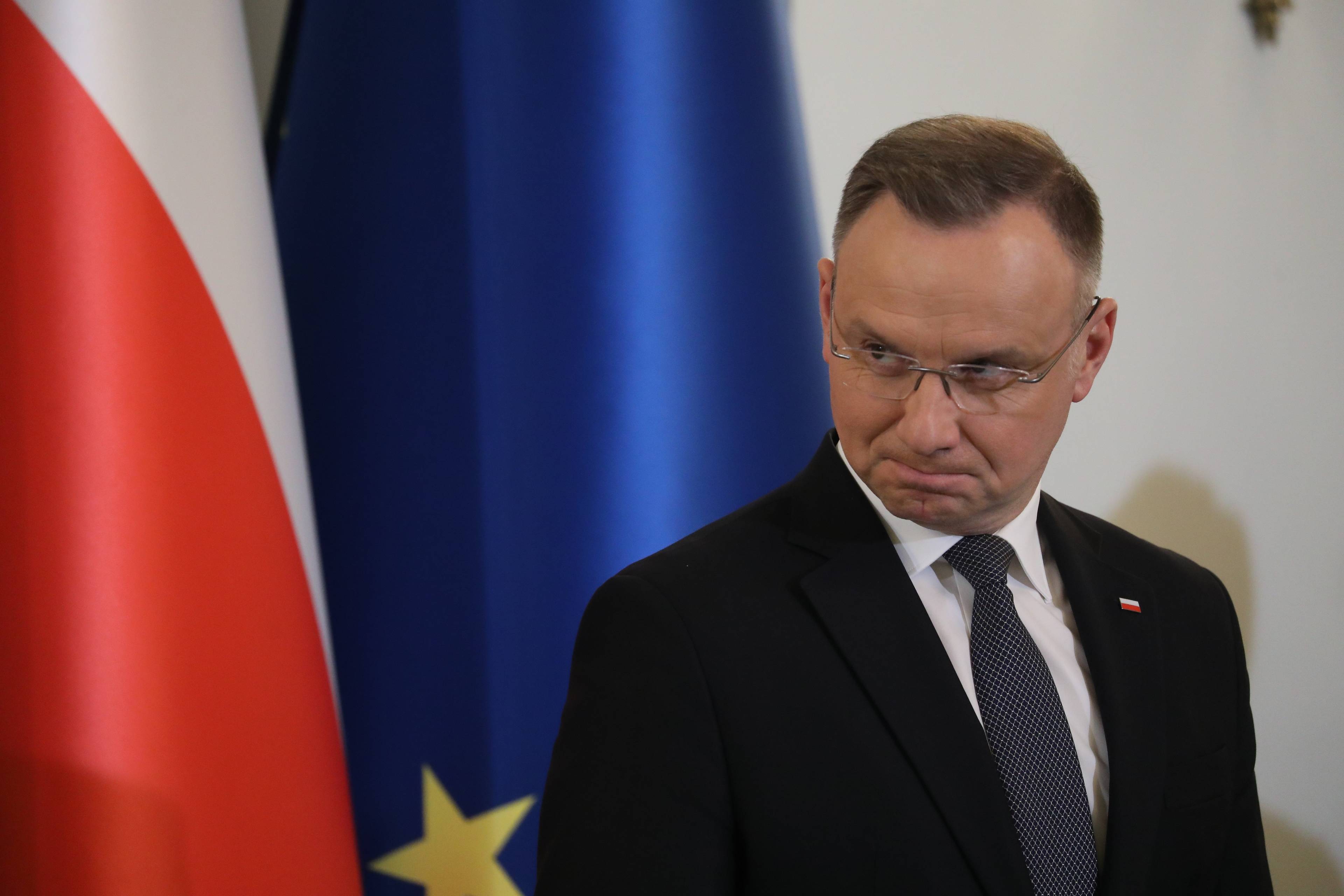 Prezydent Duda robi filuterną minę, za nim flagi Polski i Unii Europejskiej