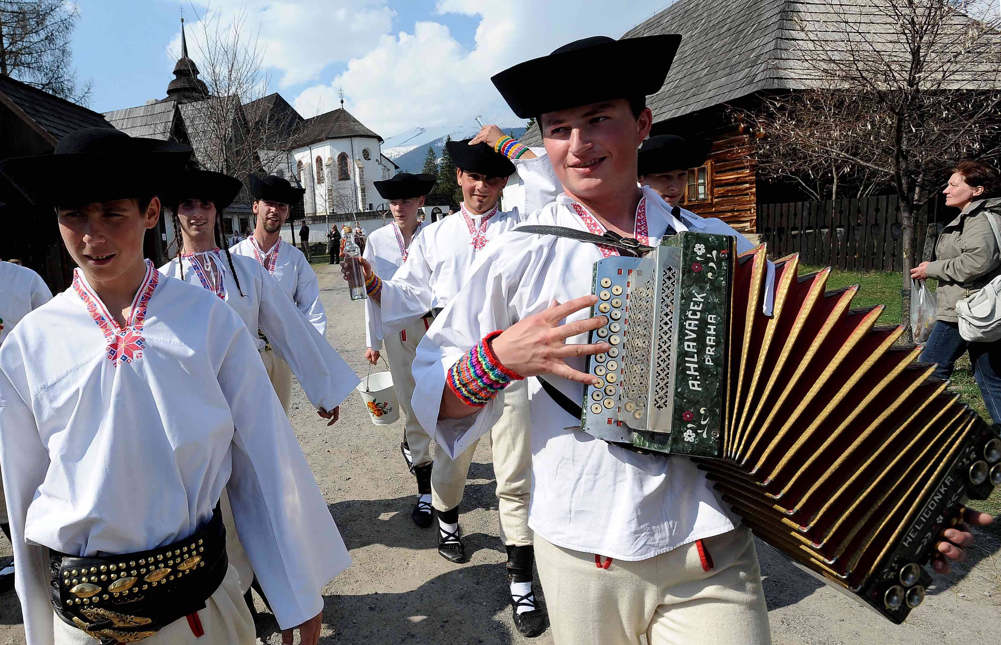 Mężczyźni w słowackich strojach ludowych idą przez wieś, jeden z nich gra na akordeonie