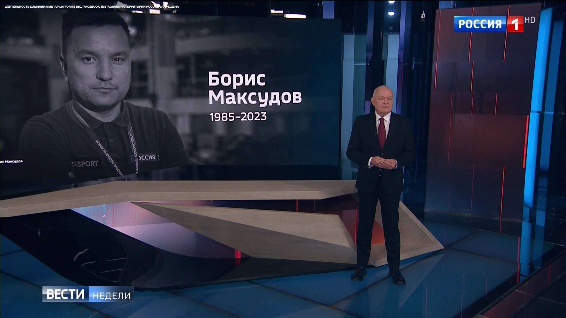 Starszy prezenter w studio. Na ekranie zdjęcie młodszego męzczyzny z rosyjskim podpisem "Borys Maksudow 1985-2023"