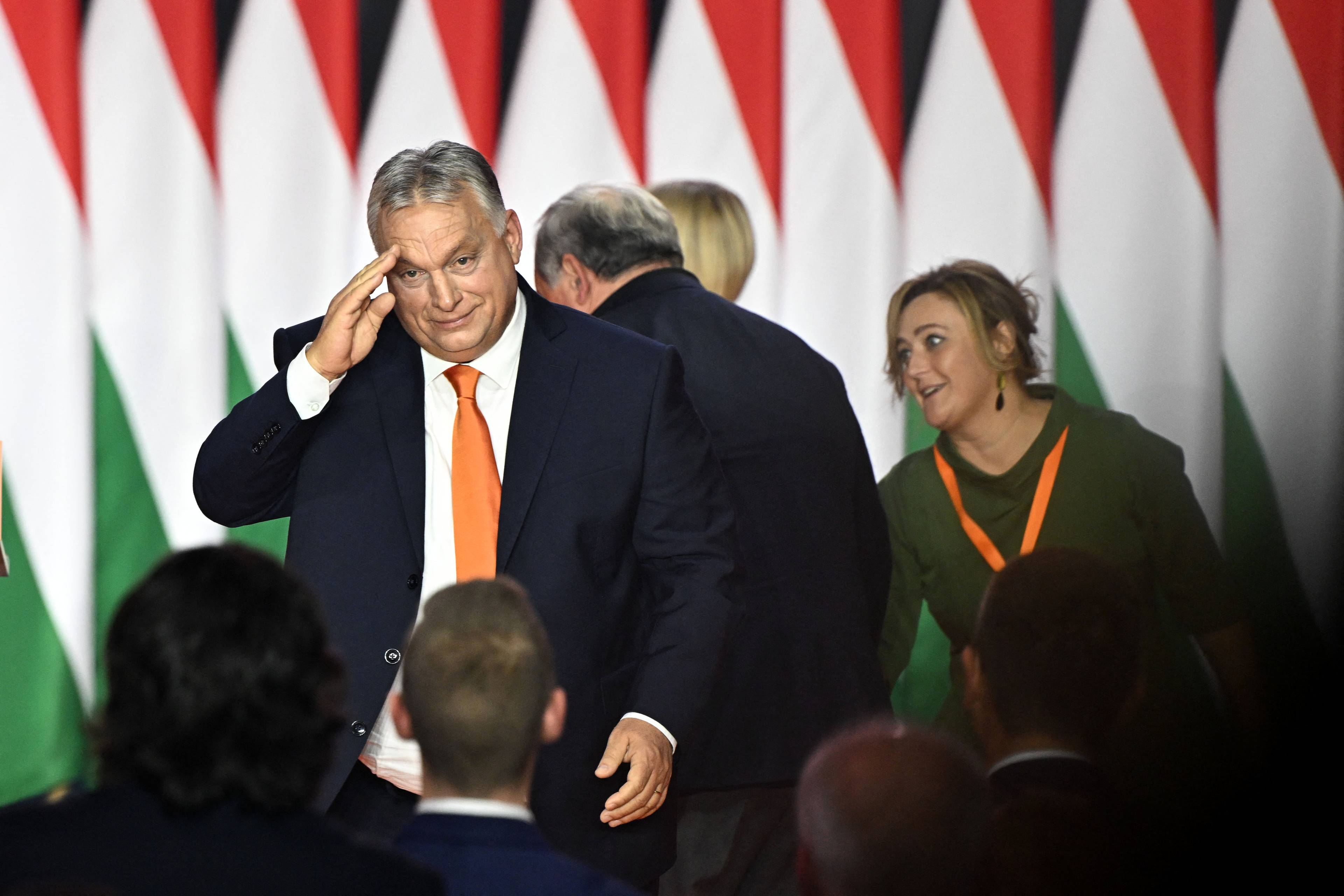 60-latek z lekką nadwagą (Viktor Orbán) podnosi rękę do czoła w geście przypominającym salut. W tle trzy inne osoby i flagi Węgier