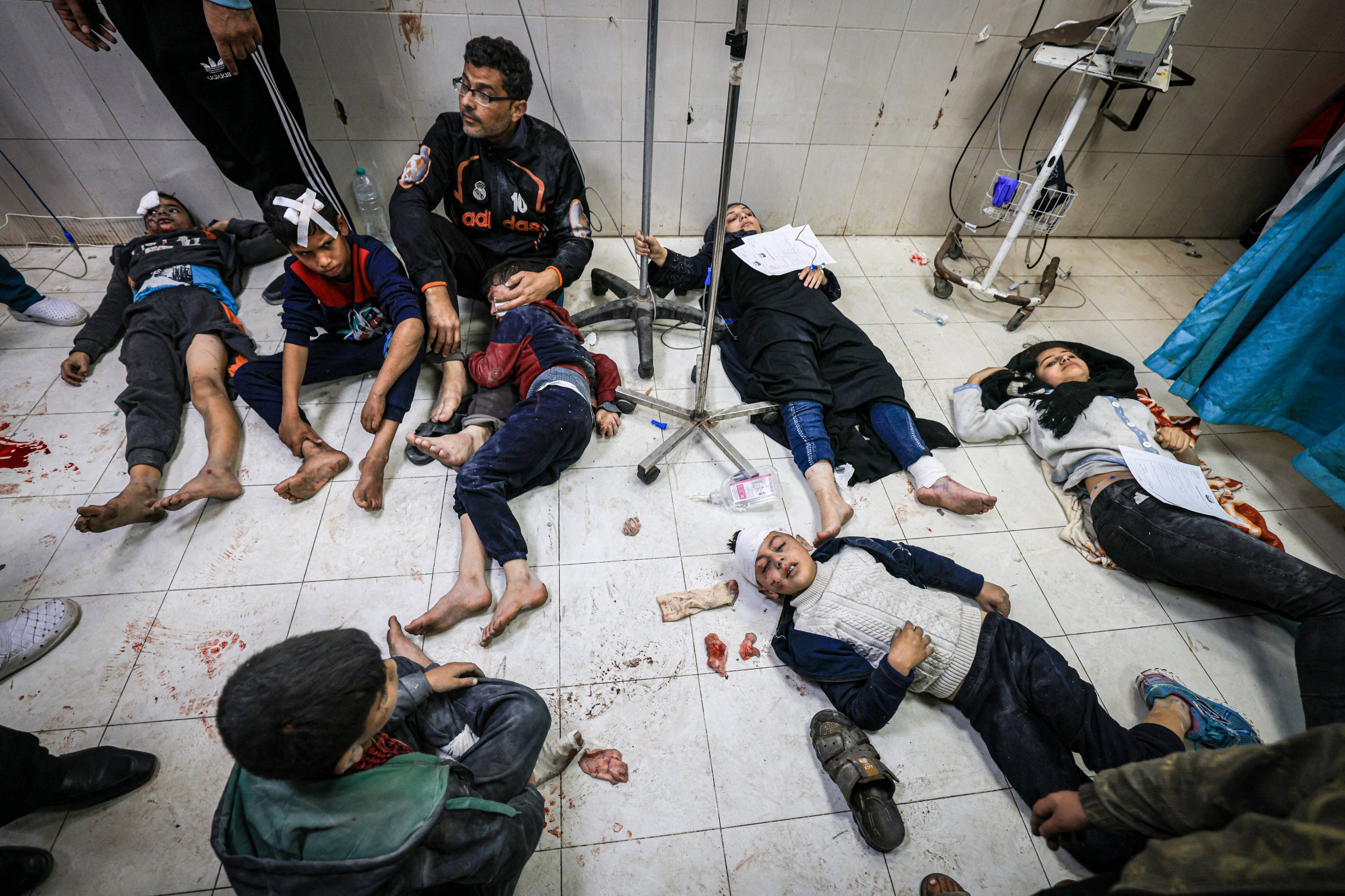 Zakrwawiony mężczyzna opierający się o ścianę, wokół leżą ranne dzieci. Szpitale w Strefie Gazy