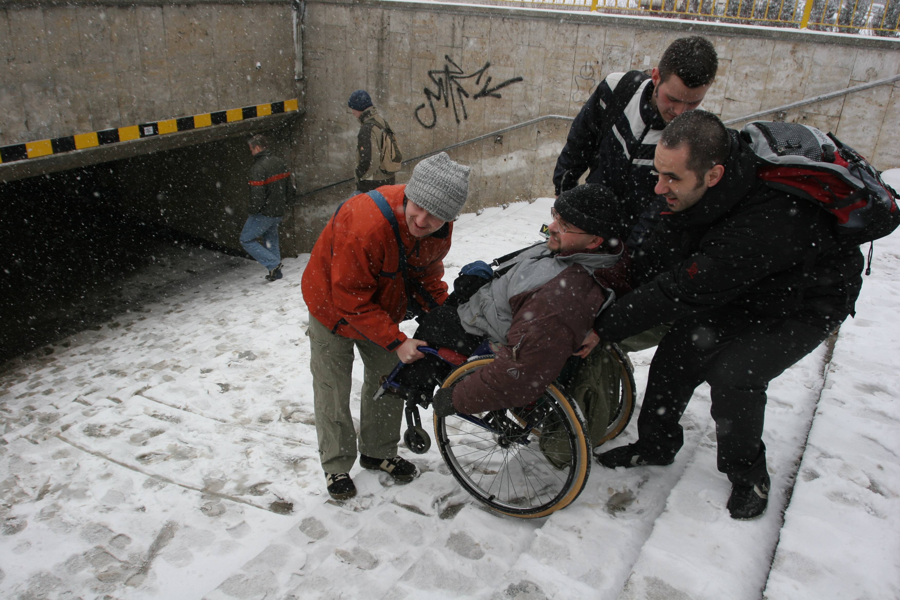 Trzej mężczyźni znoszą człowieka na wózku inwalidzkim po ośnieżonych schodach