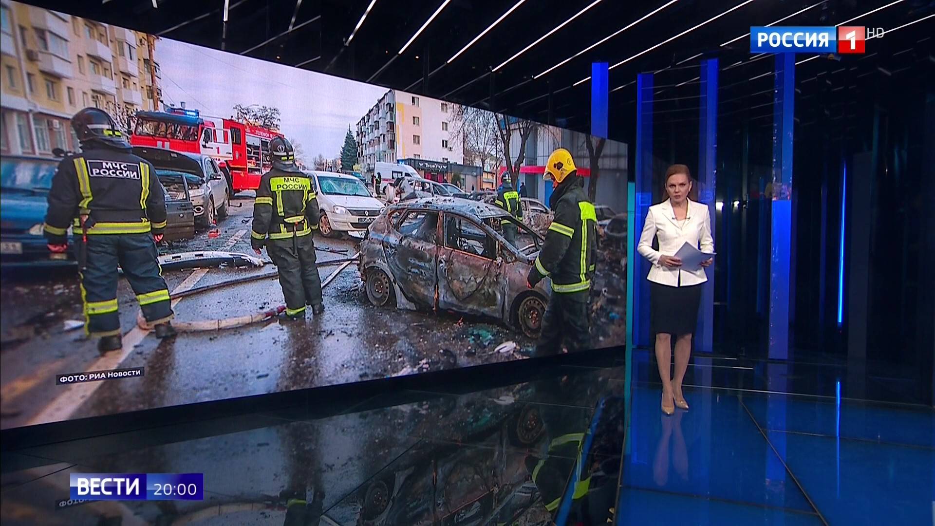 Kadr ze studia telewizyjnego. Prowadząca pokazuje ruiny miasta. Ratownicy mają kamizelki rosyjskiego ministerstwa spraw nadzwyczajnych