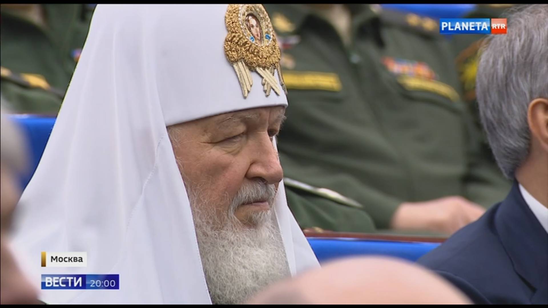 Prawoslawny duchowny w białym nakryciu głowy siedzie obok wojskowych