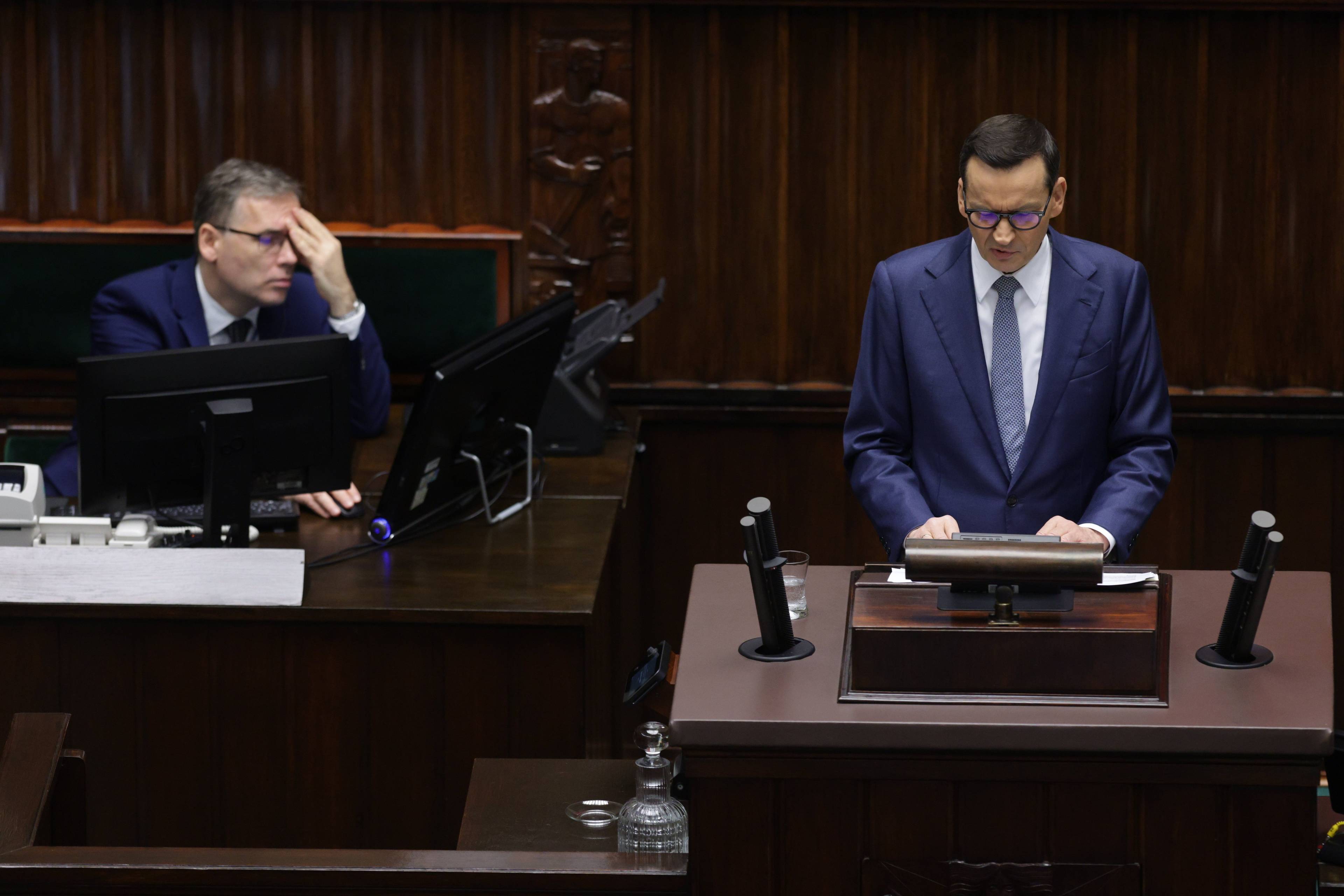 Premier Morawiecki przemawia w Sejmie, protokolant po jego lewej stronie ma zamknięte oczy i chowa twarz w dłoni