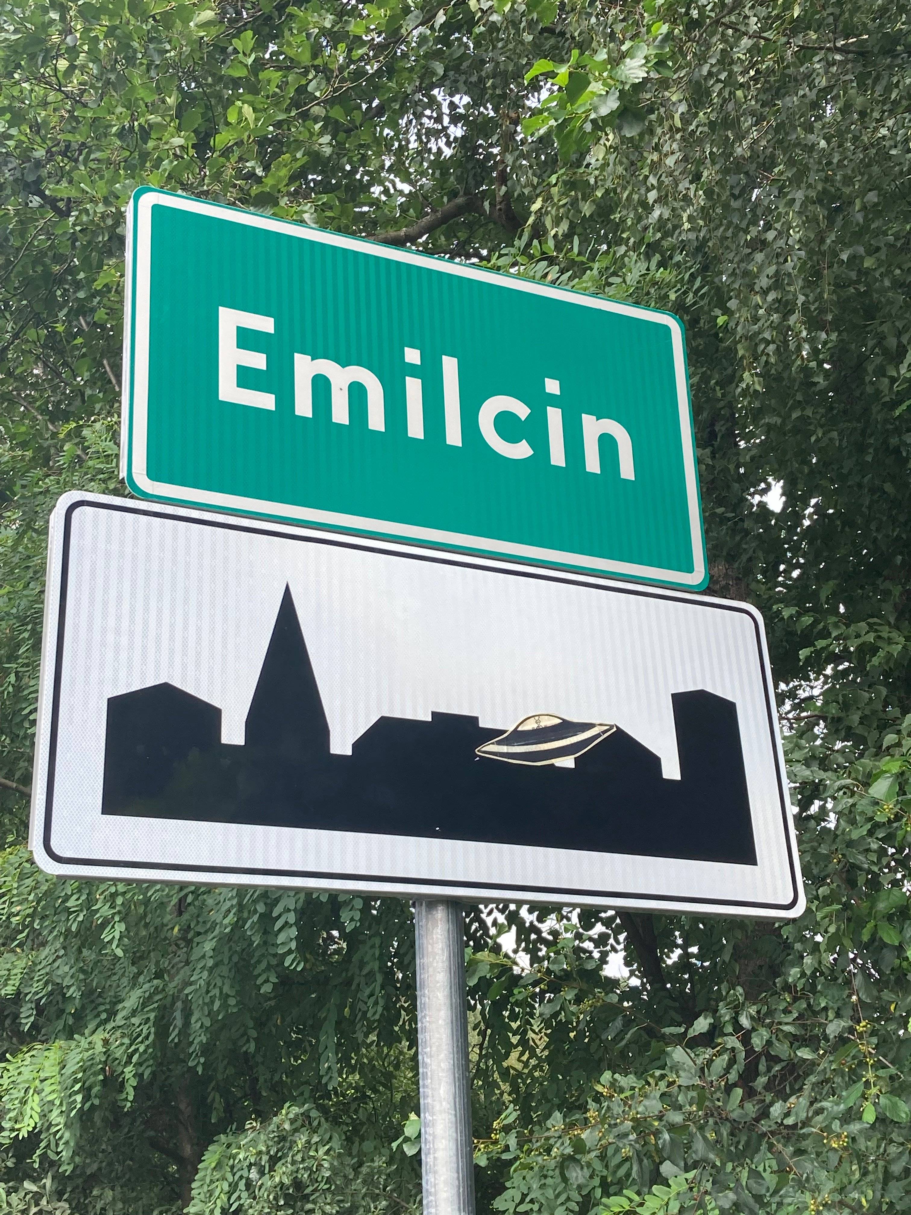 zdjęcie: zielona tablica z nazwą Emilcin, pod nią typowa biało czarna tablica oznaczająca teren zabudowany, ale nad sylwetą miasta doklejony rysunek UFO