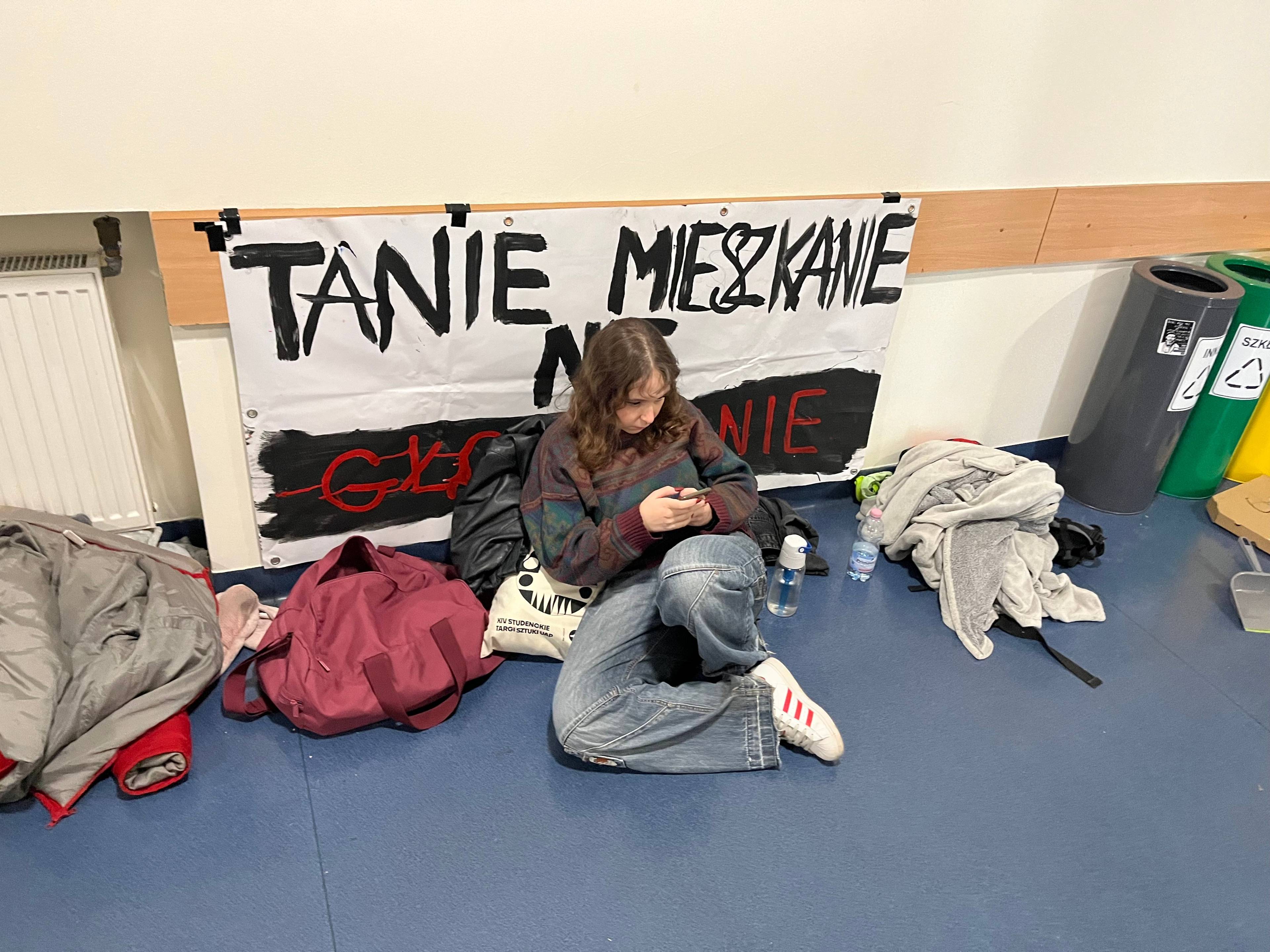 Studentka siedzi na podłodze pod plakatem okupacyjnym na korytarzu akademika