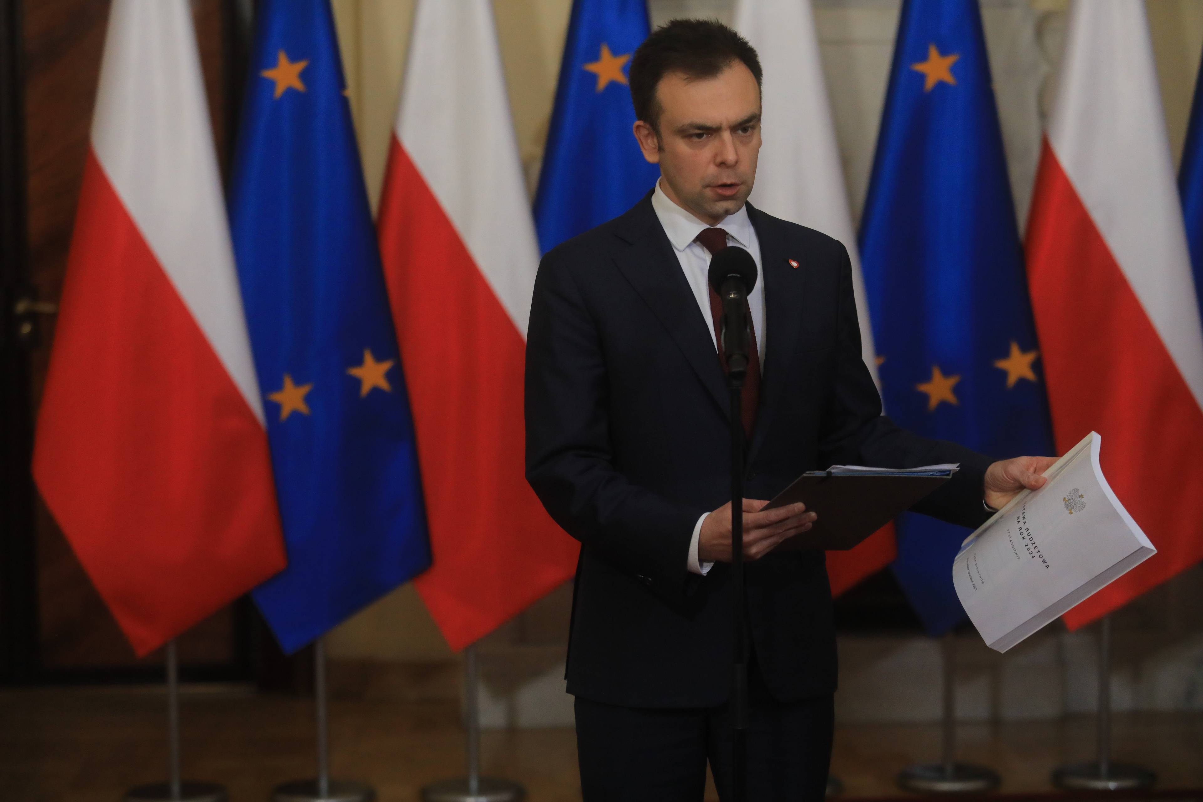 Minister finansów Andrzej Domański na konferencji prasowej stoi przed mikrofonem na tle flag UE i Polski