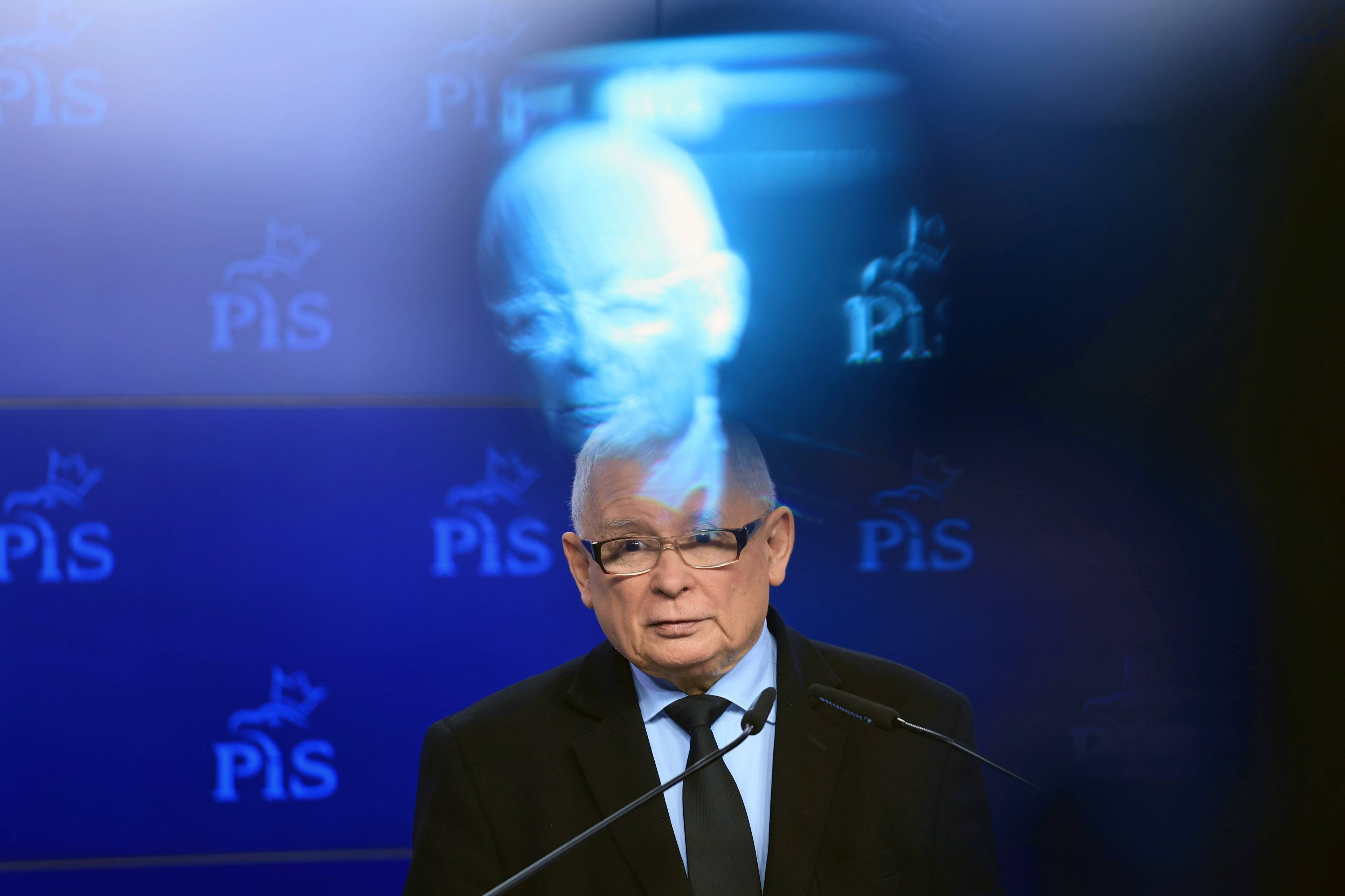 Jarosław Kaczyński przemawia, w tle ściana z logo PiS, nad politykiem efekt fotograficzny negatywu jego twarzy