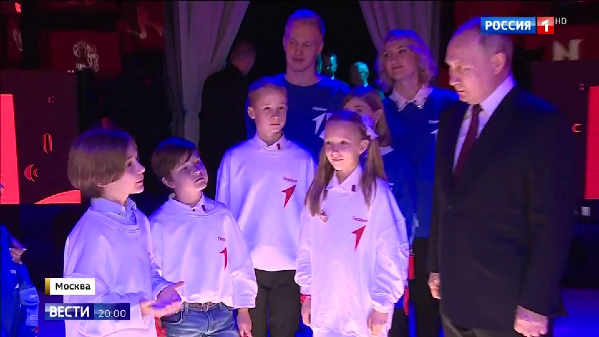 Putin i dzieci w białych bluzach. Oświetlenie sugeruje nowoczesność