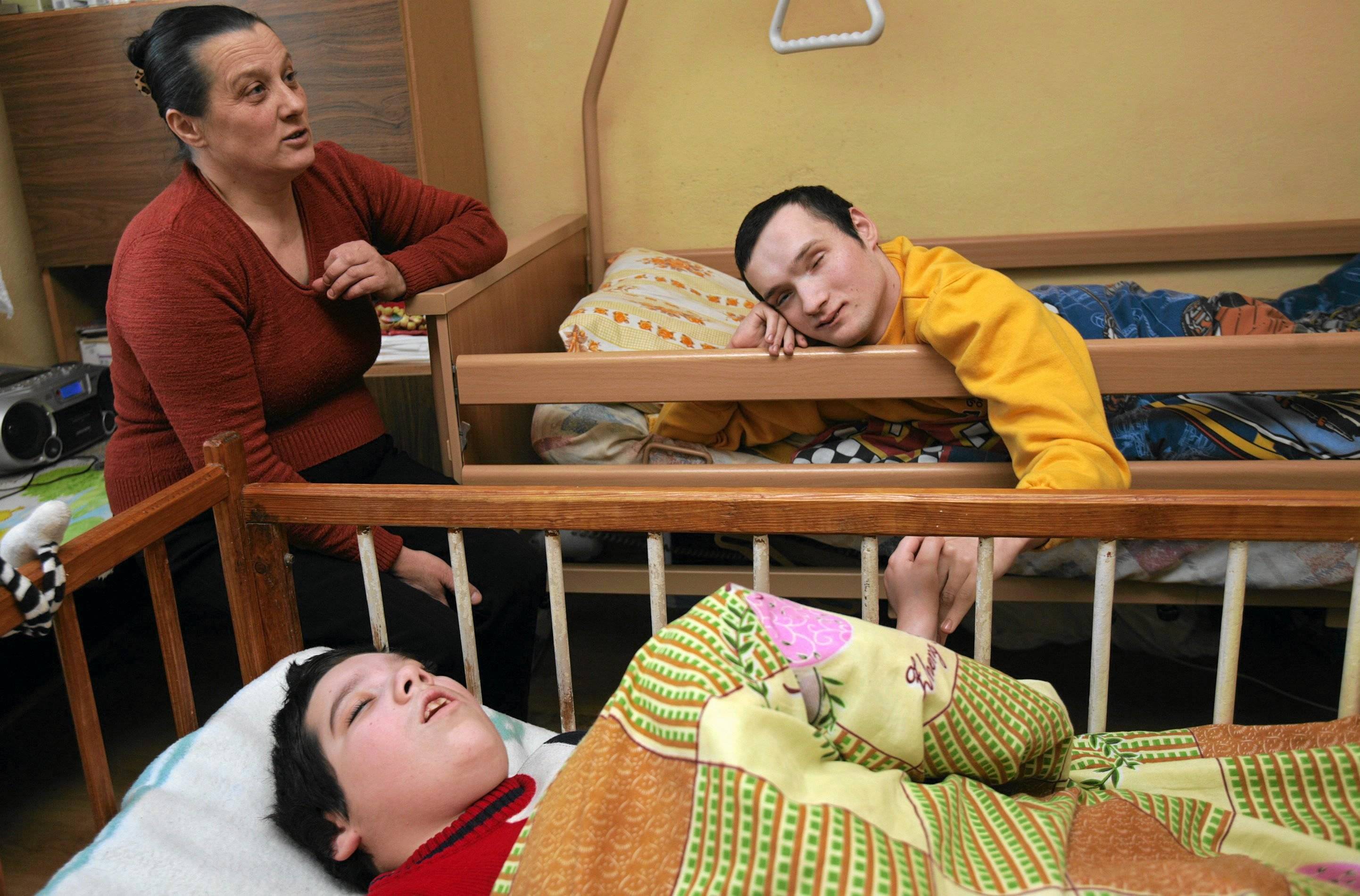 Kobieta patrzy na dwie osoby z niepełnosprawnością leżące w łóżkach