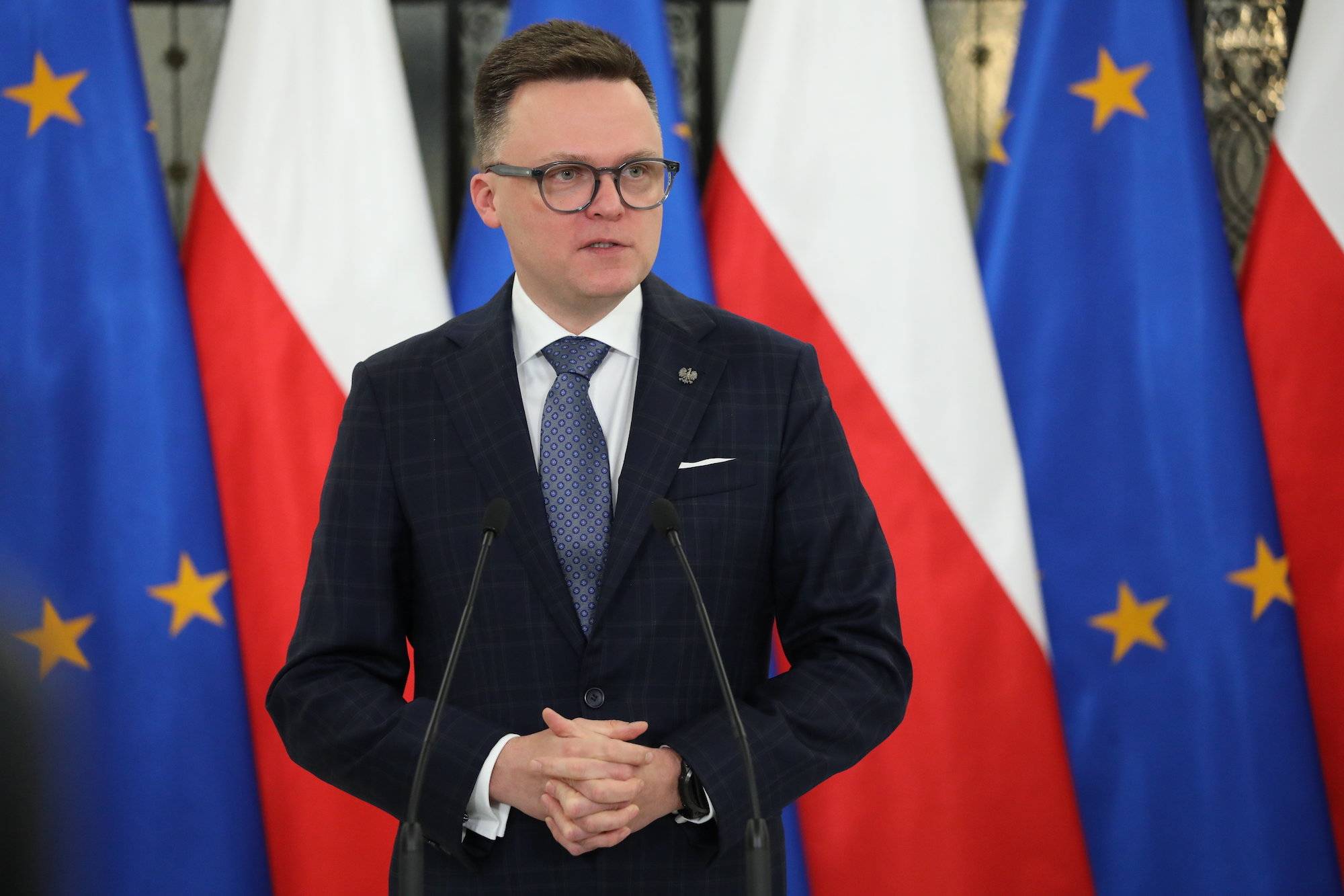 Szymon Hołownia na konferencji prasowej na tle flag Polski i Unii Europejskiej. Mówi do mikrofonów ubrany w garnitur i niebieski krawat.