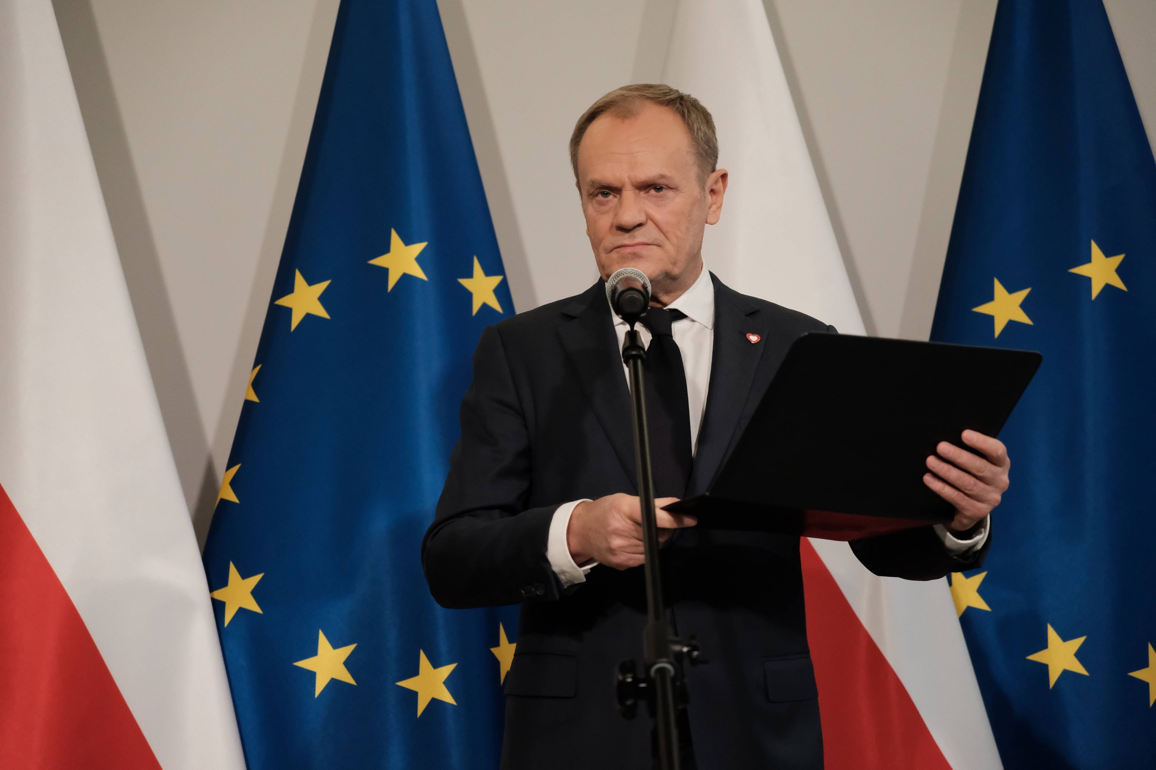 Donald Tusk na tle flag unijnych i biało-czerwonych.