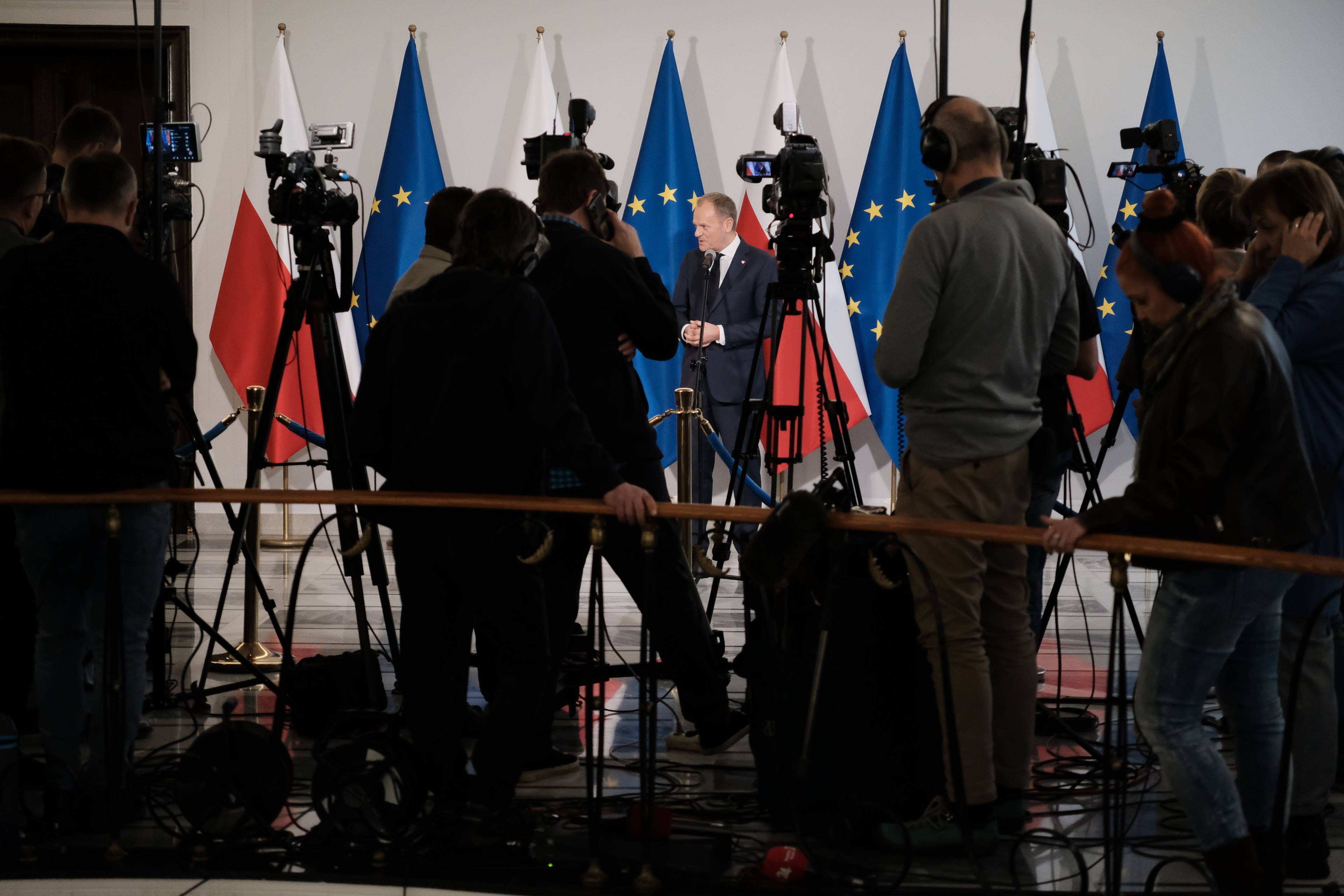 Donald Tusk przed dziennikarzami na tle polskich i unijnych flag
