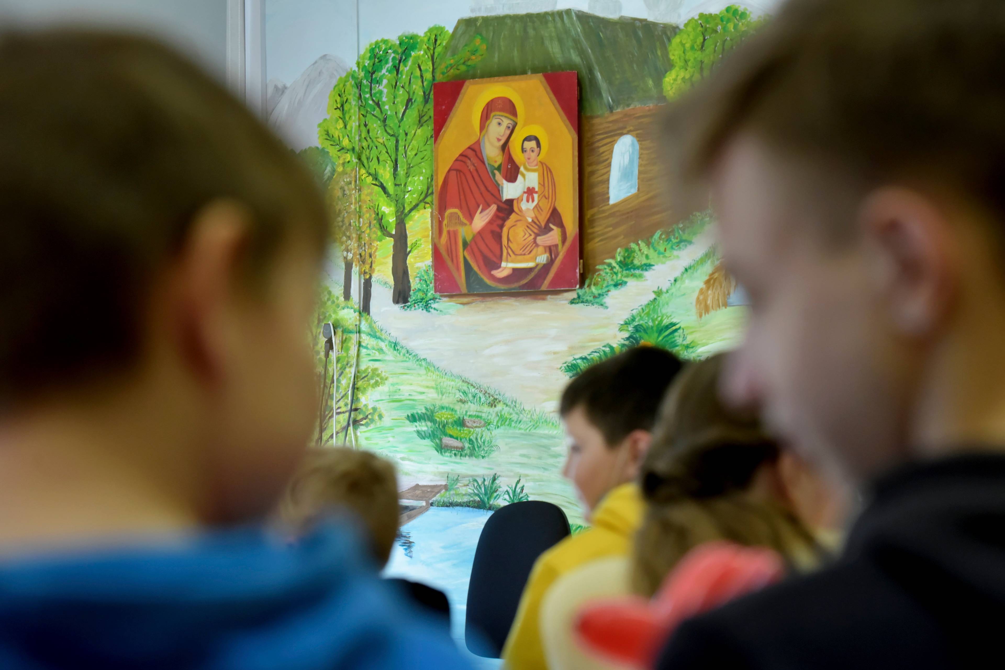 Obraz Matki Boskiej z Dzieciątkiem na ścianie szkolnej klasy. Na pierwszym planie nieostre twarze uczniów.