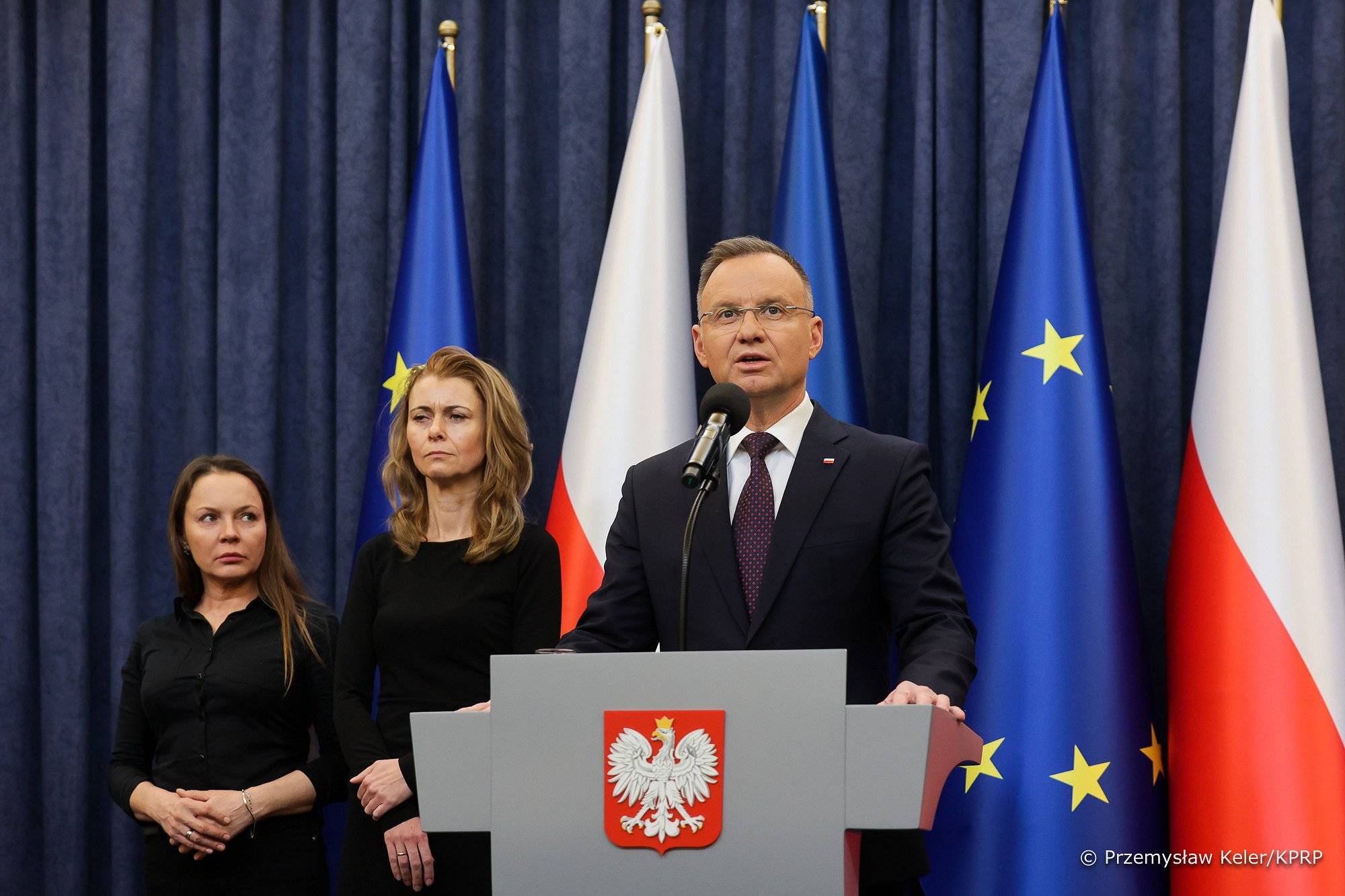 Na zdjęciu widzimy Andrzeja Dudę, za nimi stoją żony Mariusza Kamińskiego i Macieja Wąsika. W tle widzimy flagi Polski i uNII eUROPEJSKIEJ