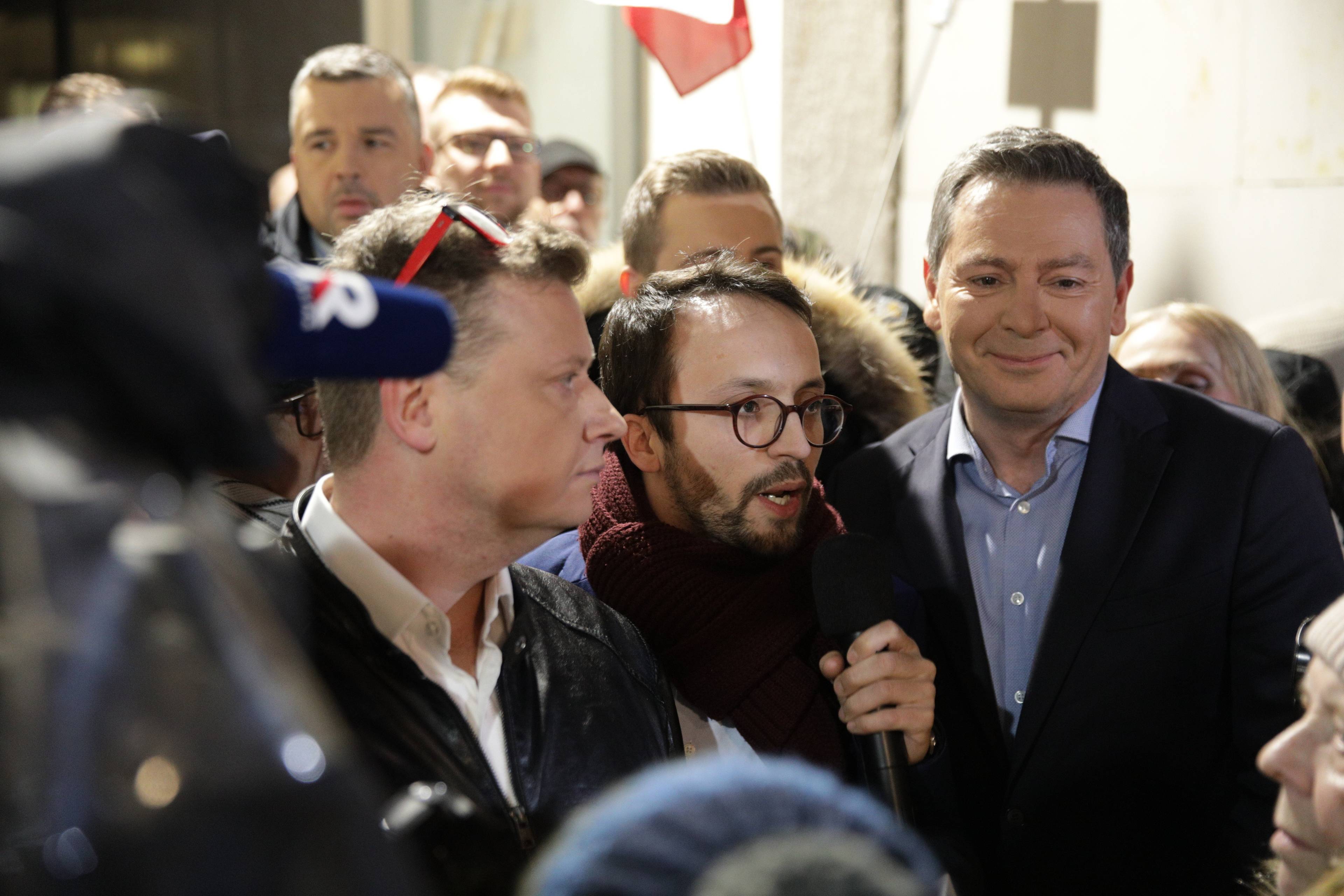 Marcin Tulicki (z okularami w czerwonych oprawkach na głowie), Samuel Pereira (z mikrofonem) i Michał Adamczyk (uśmiecha się) podczas manifestacji pod siedzibą TVP. Z tyłu Michał Rachoń