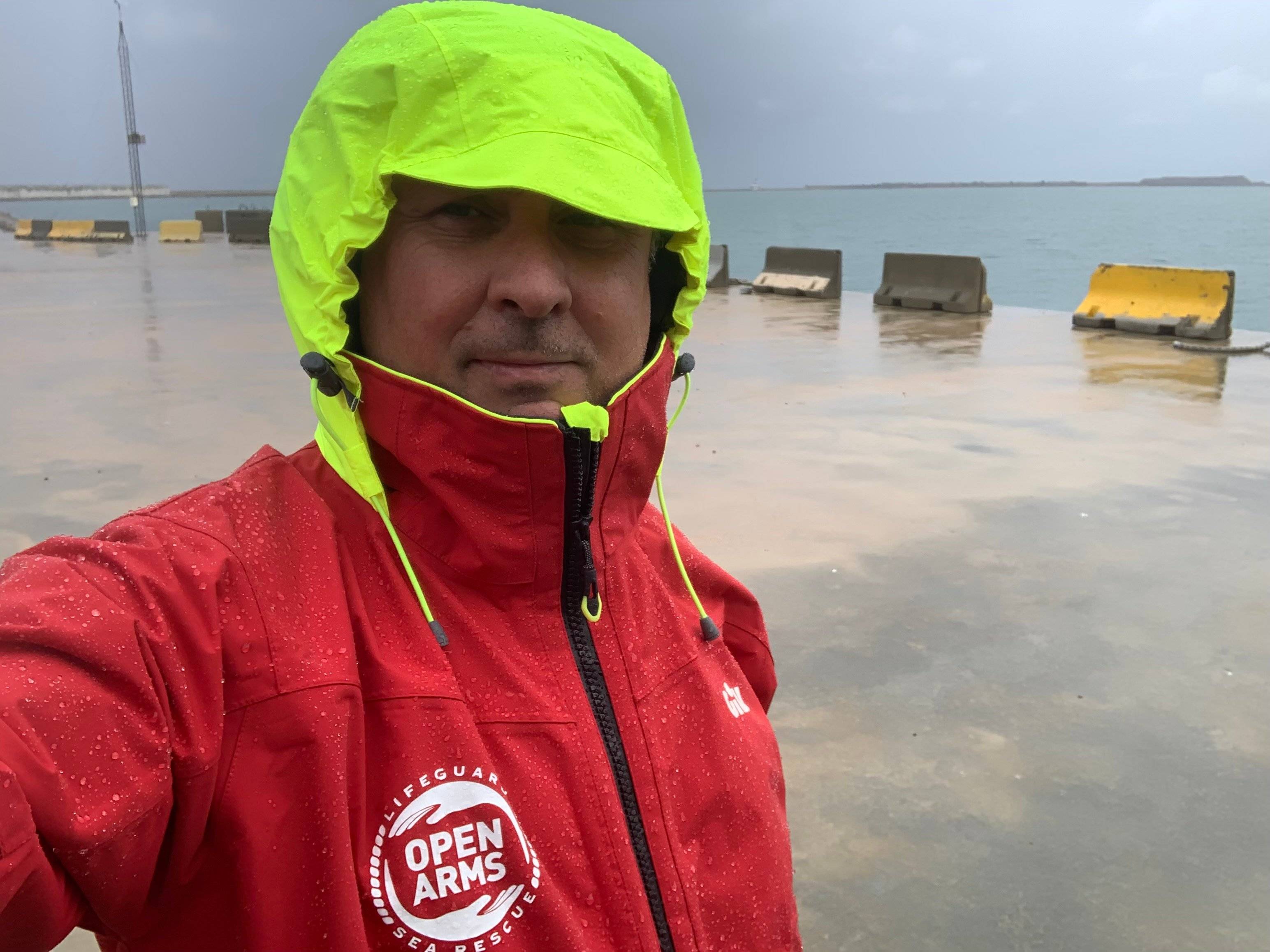 mężczyzna w czerwonej kurtce z żółtym kapturem nasuniętym na czoło, w głębi betonowe nabrzeże i morze