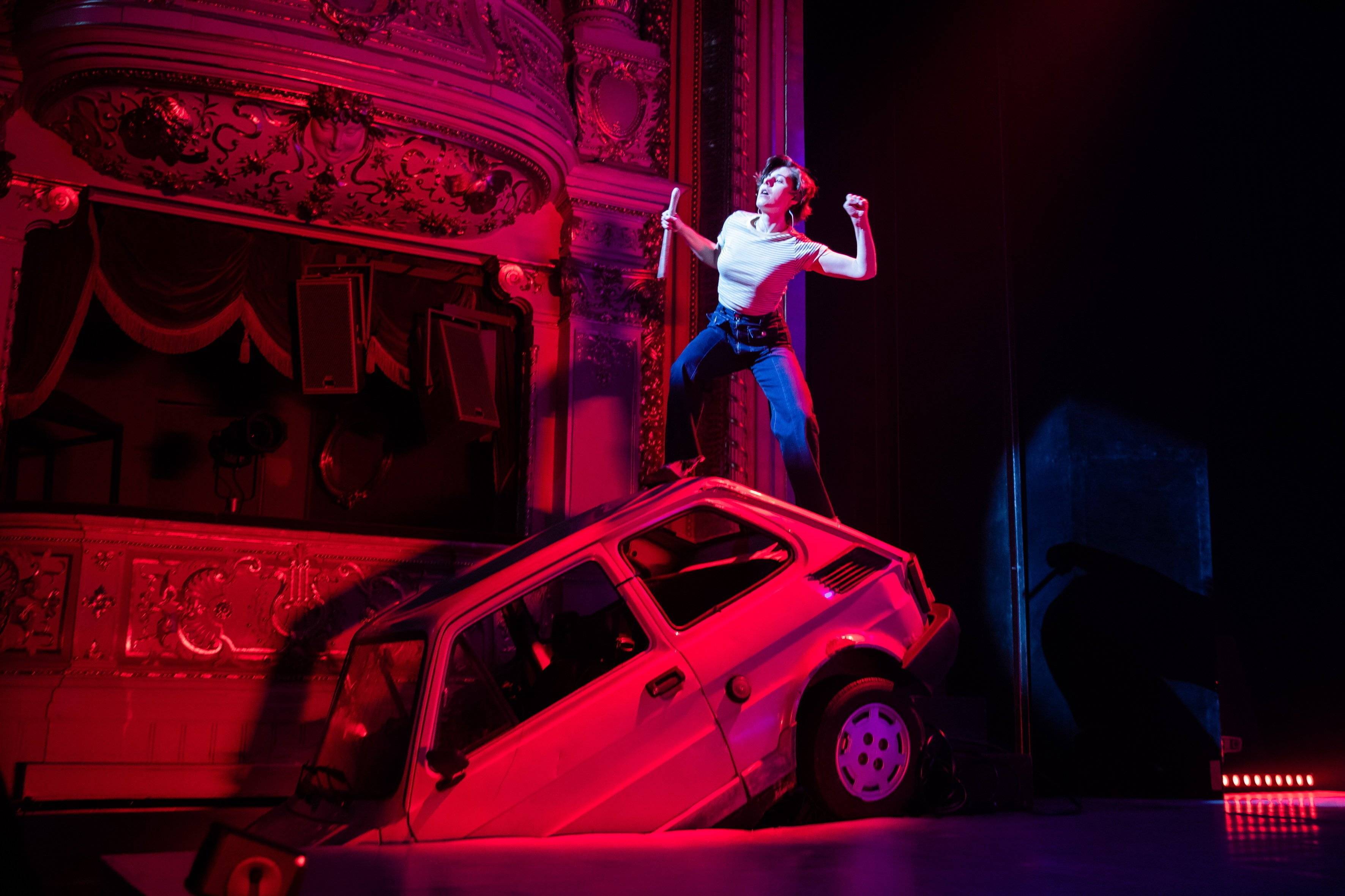Aktorka stoi na przewróconym małym fiacie, który jest elementem scenografii teatralnej, w tle widać loże teatralne w czerwonym świetle. Musical 1989. Teatr TV