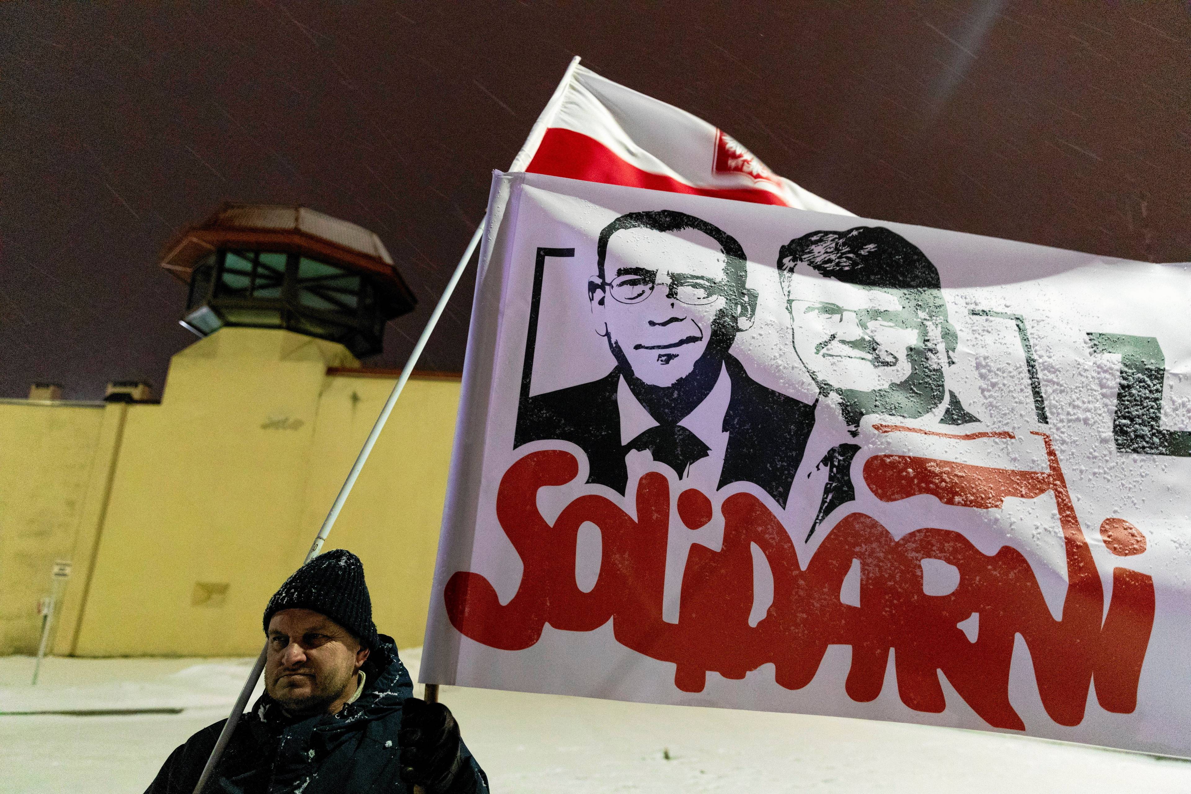 Mężczyzna trzyma transparent z czarno-białymi wizerunkami Mariusza Kamińskiego i Macieja Wąsika i czerwonym napisem Solidarni. W tle mur więzienia w Radomiu