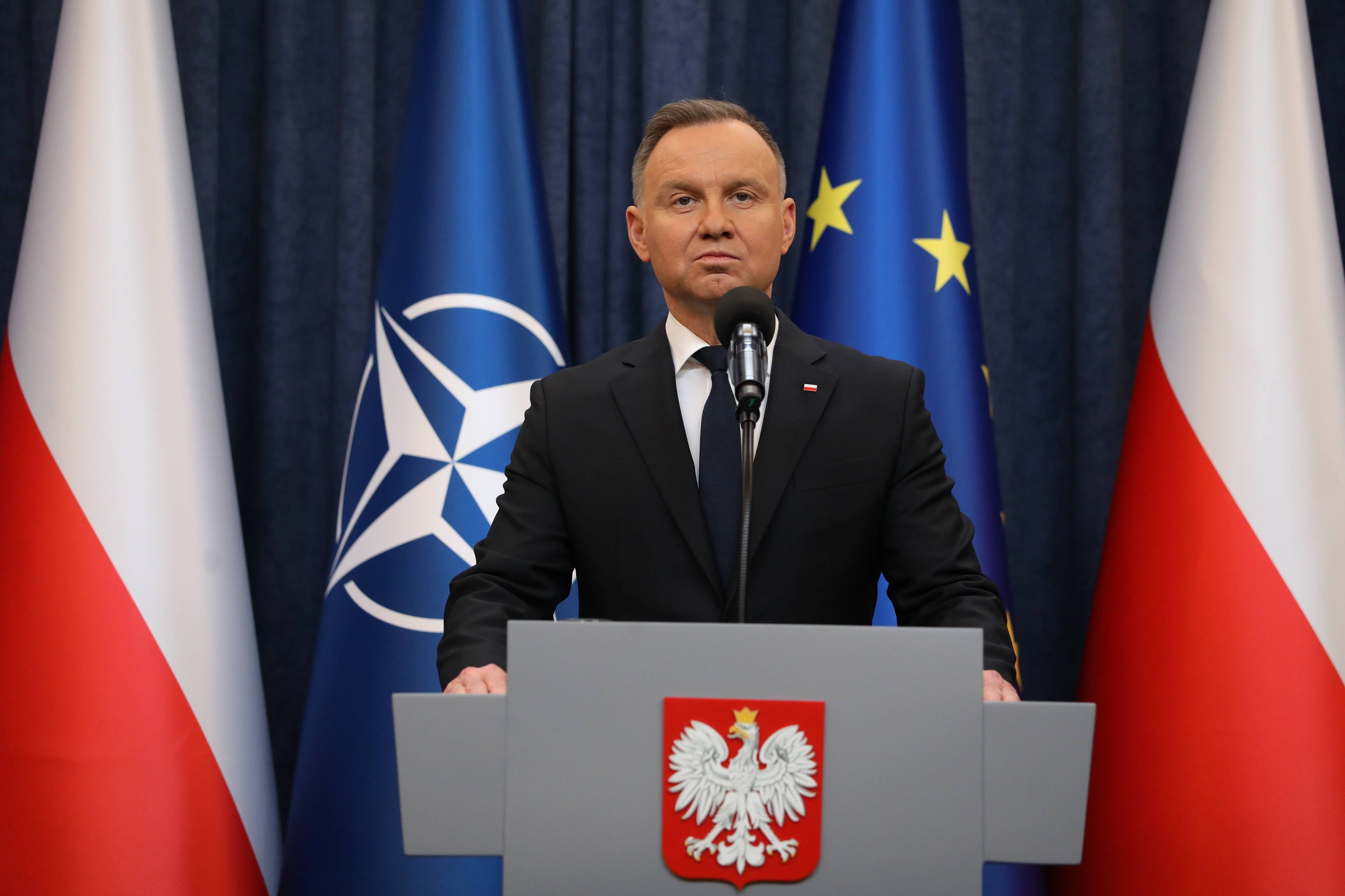 Andrzej Duda z poważną miną za mównicą, w tle flagi NATO, Unii Europejskiej i Polski