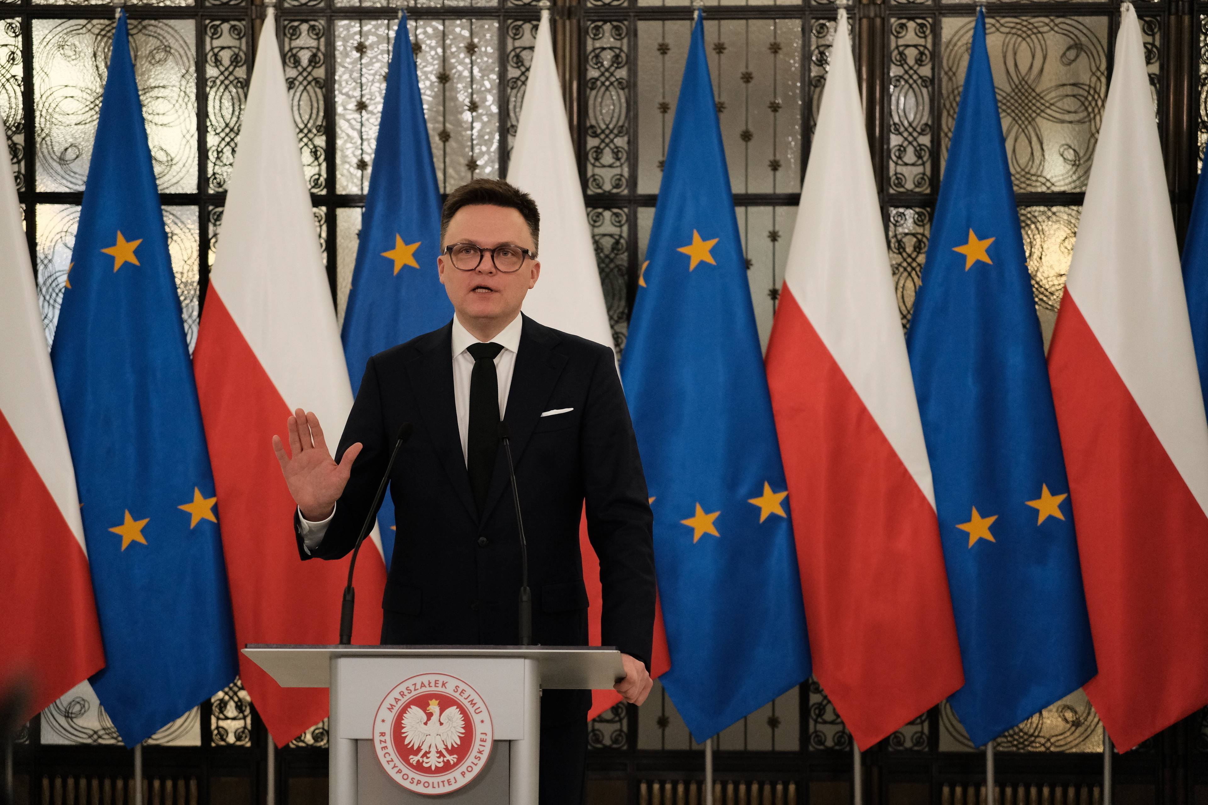40-latek w okularach i garniturze przemawia na tle flag polskich i unijnych