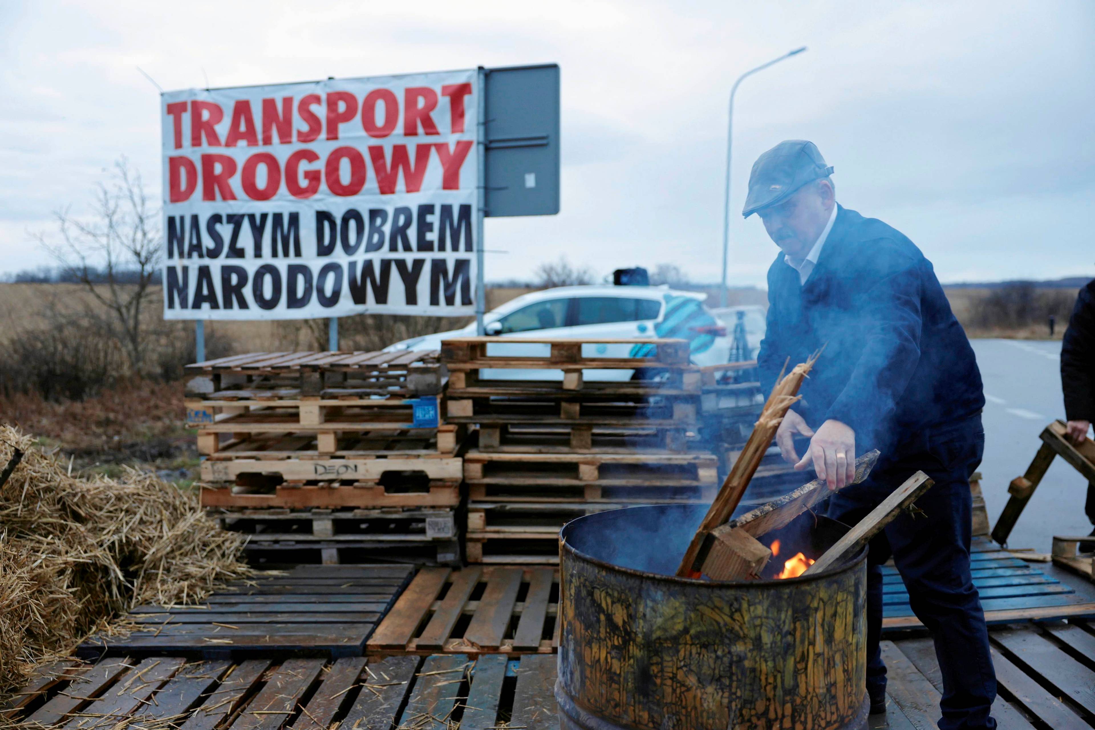 Protest rolników na granicy, mężczyzna wrzuca deski do metalowego kosza, w którym płonie ogień. w tle transparent z hasłem „Transport drogowy naszym dobrem narodowym”
