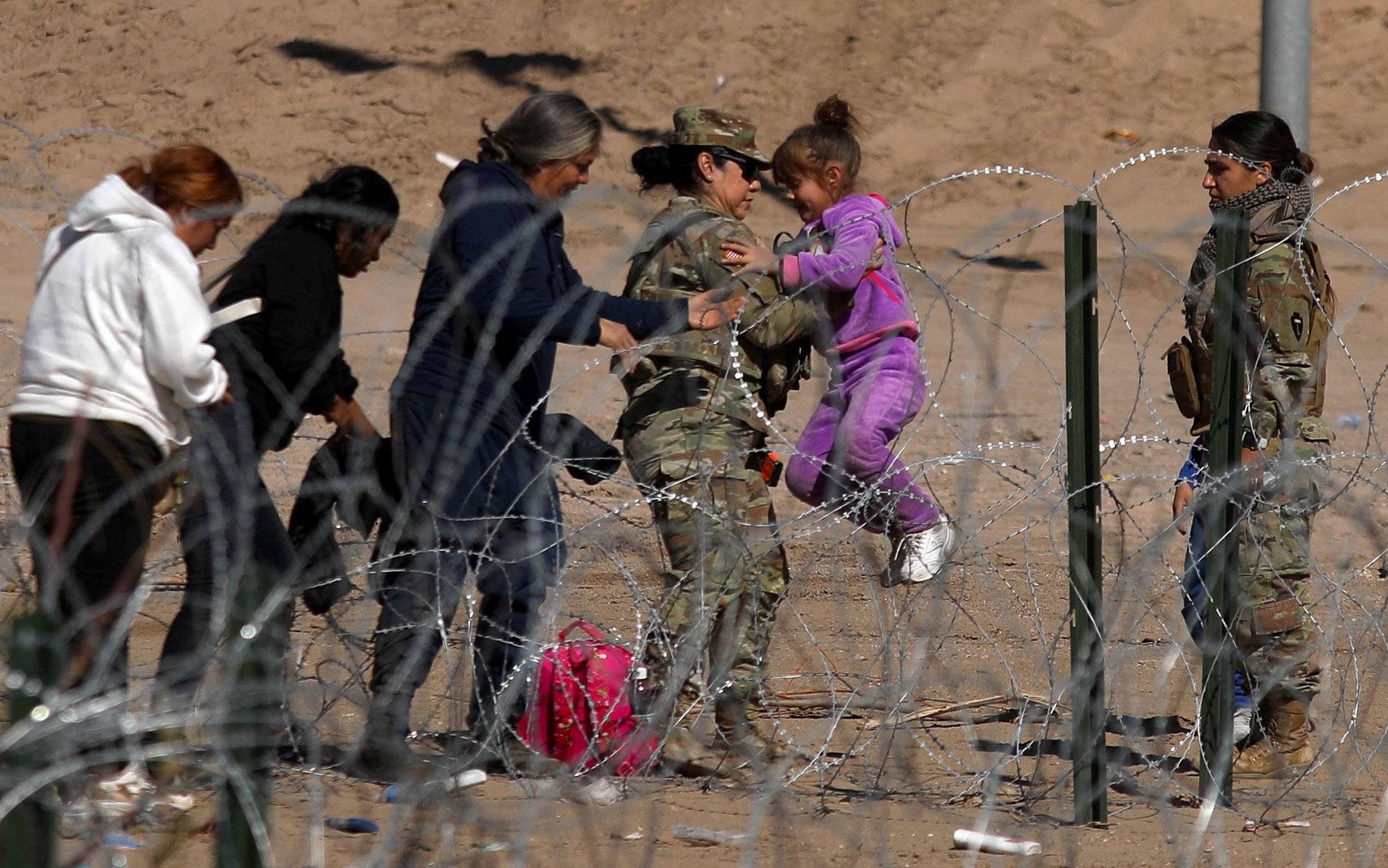 Strażniczka graniczna trzyma dziecko, na ziemi leży plecak, na pierwszym planie drut kolczasty, wokół nich podłoże z piasku